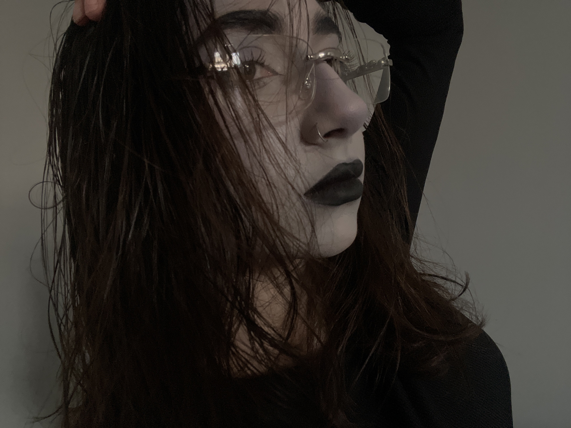 The image displays a close-up, mid-profile portrait of an individual, likely a young adult, facing towards the right. The subject's skin exhibits a pale complexion, accented by dark, possibly wet or heavily textured brown hair that partially obscures the left side of the face. The individual wears thin-framed glasses, whose lenses reflect light, and has dark, pronounced eyebrow makeup and dark eye makeup. The lips are covered in a matte, dark, possibly black, lipstick. A silver-toned ring is visible piercing the left nostril. The subject's right arm is partially visible at the top of the frame, with the hand positioned near the back of the head or in the hair. The individual is wearing a dark, long-sleeved garment of a textured fabric. The background is a plain, light grey wall, indicating an indoor setting. No specific geographical features or identifying markers related to the city of Ternopil, Ukraine, are discernible within the visible elements of the scene.