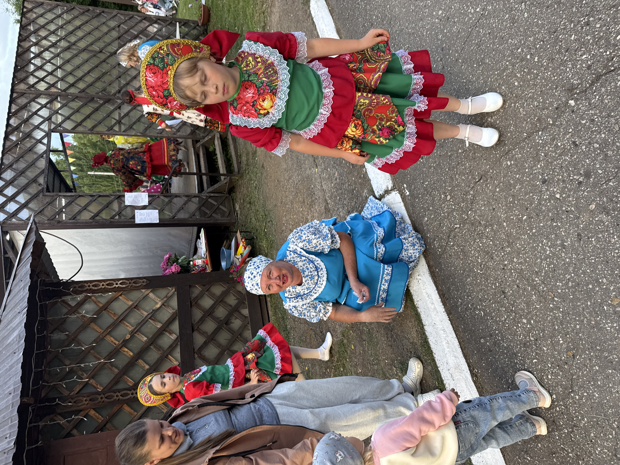 This image captures a scene in the city of Kukuya, Russia, during an outdoor event. In the foreground, a young girl in traditional red and green Russian attire, complete with a decorative headdress, stands looking to her right. To her left, a woman in a blue dress and headscarf kneels on the ground. Behind them, another young girl in similar festive clothing is partially visible. Further back, through an open wooden structure resembling a gazebo or pavilion, other individuals in traditional dress can be seen, along with decorative flags. The ground is asphalt, with a white line running across it. The background shows a building with wooden lattice work and some greenery, suggesting an outdoor cultural gathering or festival.