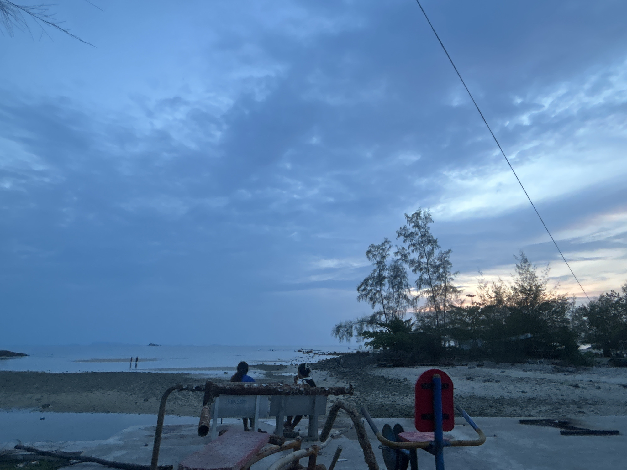 The image shows a beach scene at dusk in ตำบลเกาะพะงัน, Thailand. The sky is predominantly cloudy with a hint of sunset on the horizon. In the foreground, there is rusty outdoor exercise equipment. Two people are seated on a concrete structure behind the exercise equipment, facing the ocean. Two additional individuals are seen walking on a sandbar in the distance. The beach consists of sand, rocks, and scattered debris. Trees line the shore, partially obscuring the horizon. A power line is visible stretching across the upper right of the sky. The overall scene conveys a calm, overcast evening at the beach.