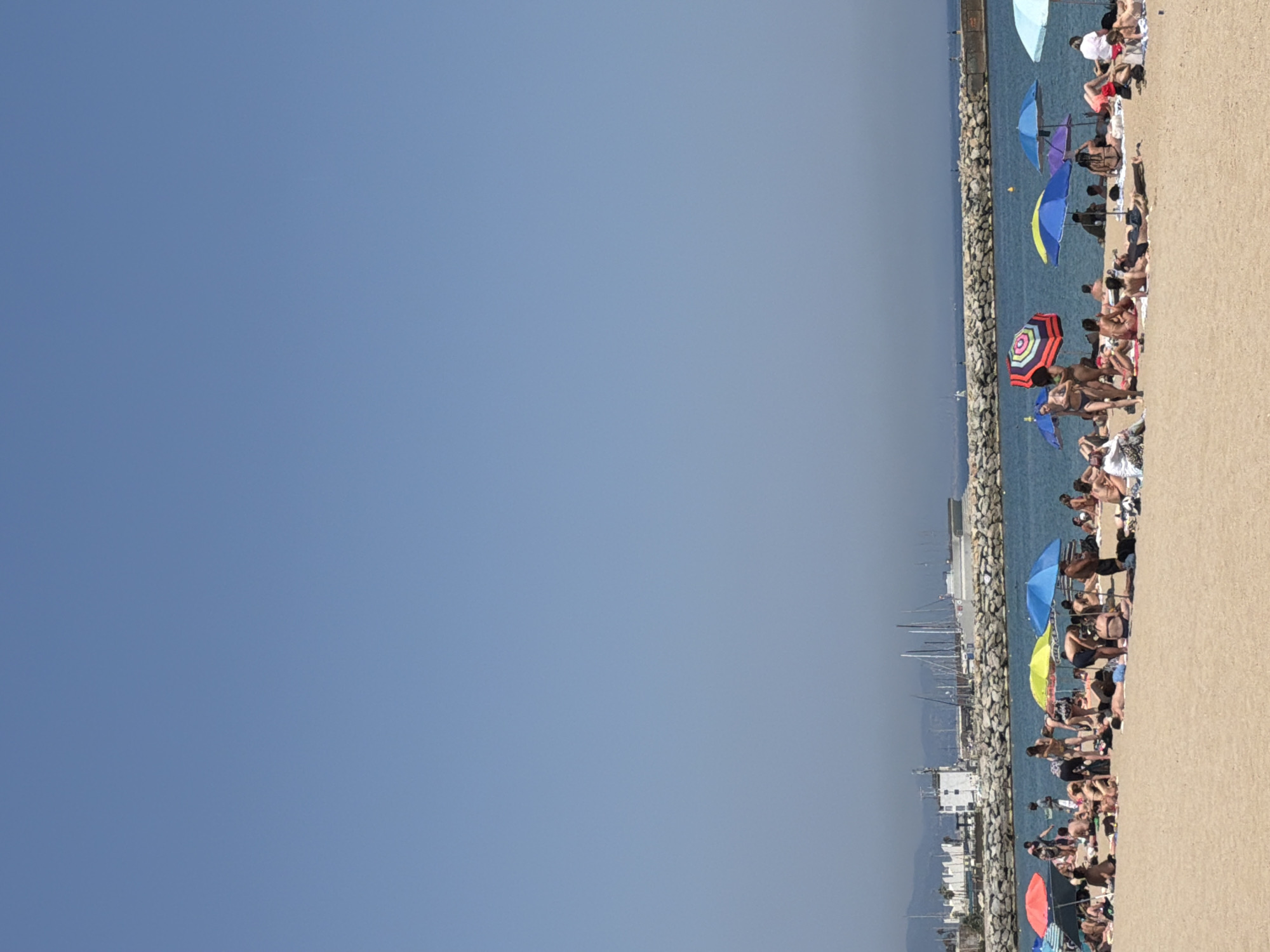The image shows a beach scene in Barcelona, Spain. The beach is crowded with people sunbathing and relaxing under umbrellas. The beach is sandy and there is a rocky breakwater separating it from the sea. In the background, there are sailboats and buildings. The sky is clear and blue.