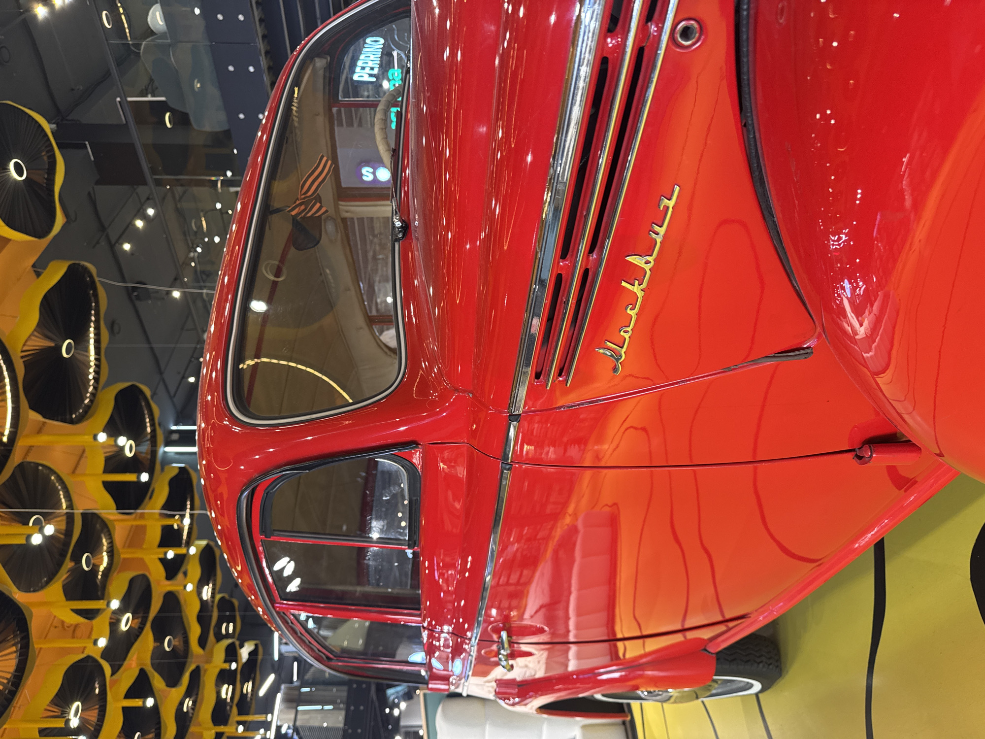 A red Moskvitch car is exhibited in Moscow, Russia. The car is positioned indoors, possibly in a museum or exhibition hall. The background features yellow and black circular decorative elements on a wall. The name "Moskvitch" is visible in gold lettering on the side of the car.
