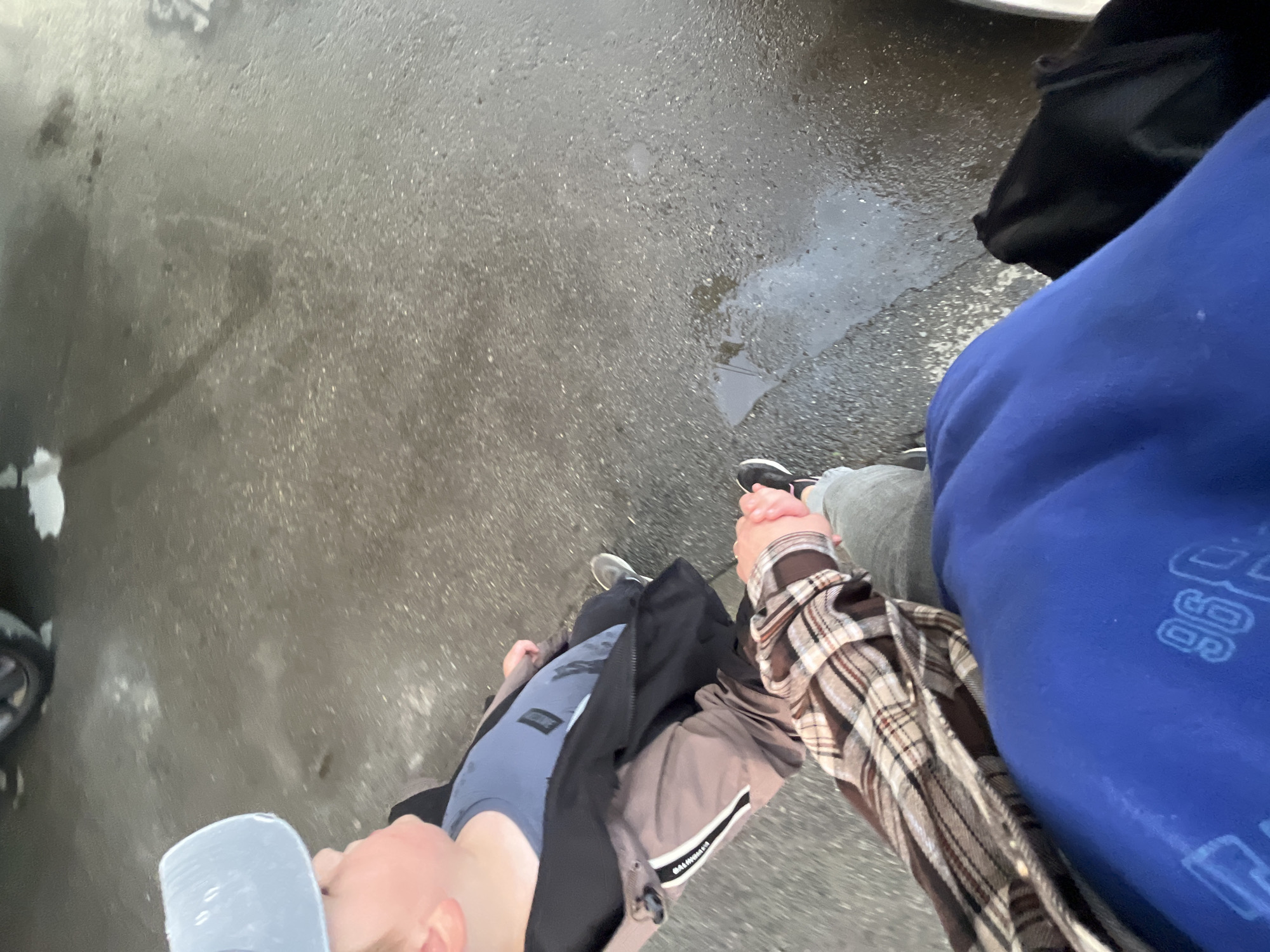 The image shows two people holding hands and walking on a wet asphalt surface, possibly during a rainy or recently rained-on day. The ground is dark and reflects light in puddles, suggesting a damp environment. One person is wearing a blue hoodie with a white graphic design on the sleeve, and the other is wearing a plaid shirt over a grey jacket and a blue t-shirt. Both are wearing athletic shoes. The perspective is from above, looking down at the subjects and the ground. The scene appears to be outdoors, likely on a street or sidewalk in Chelyabinsk, Russia. The overcast lighting and wet ground suggest it is daytime, but the exact time is not discernible. The overall mood is neutral, with the focus on the act of walking and holding hands. There is no discernible text in the image.