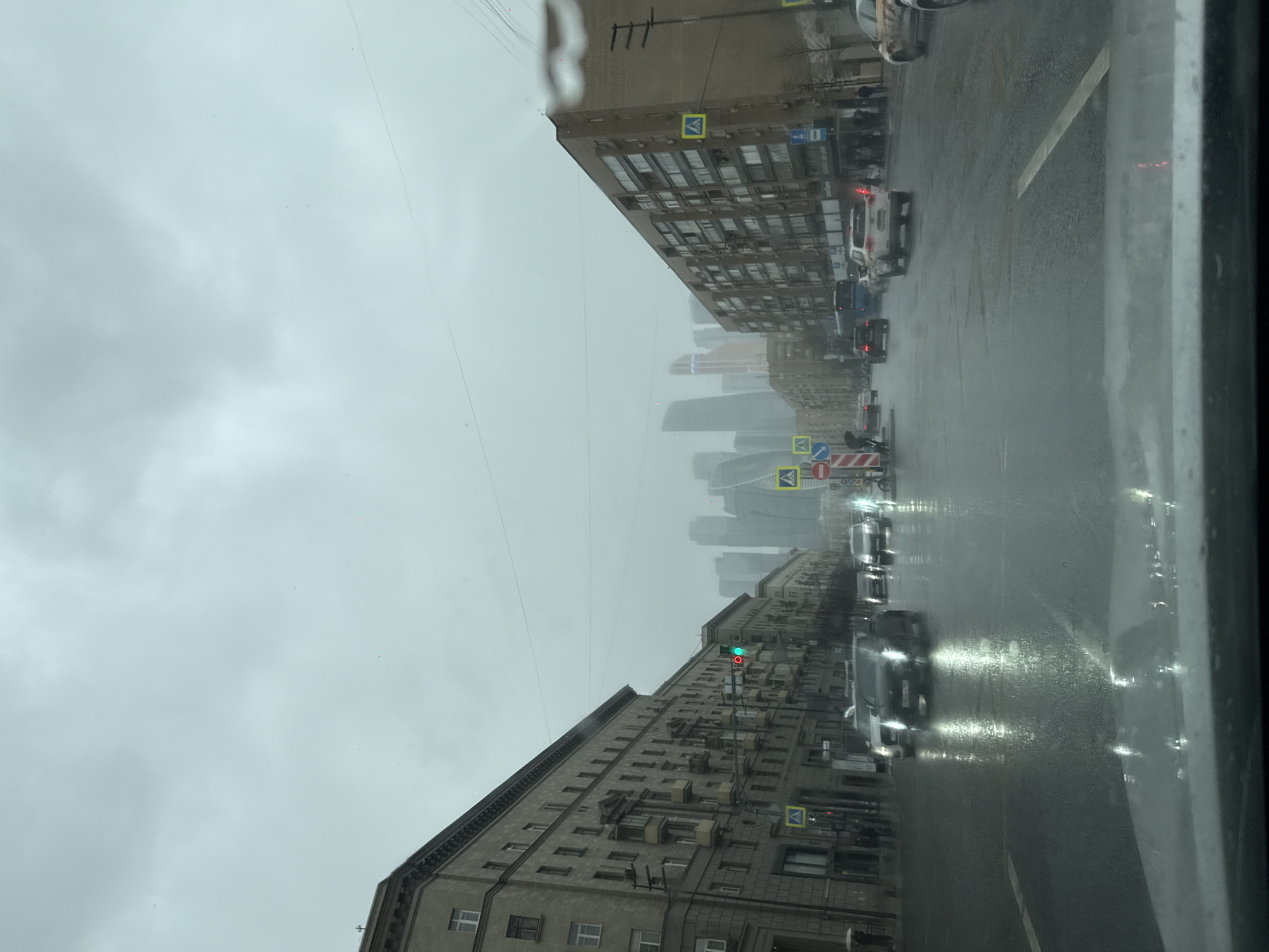 The image shows a rainy street scene in Moscow, Russia, taken from inside a vehicle. The view is oriented down a straight avenue flanked by tall, multi-story buildings on either side. In the distance, the modern skyscrapers of the Moscow International Business Center (Moscow City) are visible through the mist and low-hanging clouds. The street is wet and reflects the headlights of the moving vehicles. Pedestrian crossings are marked by signage. A traffic light is visible, indicating a green light. The overall atmosphere suggests a cold and overcast day.