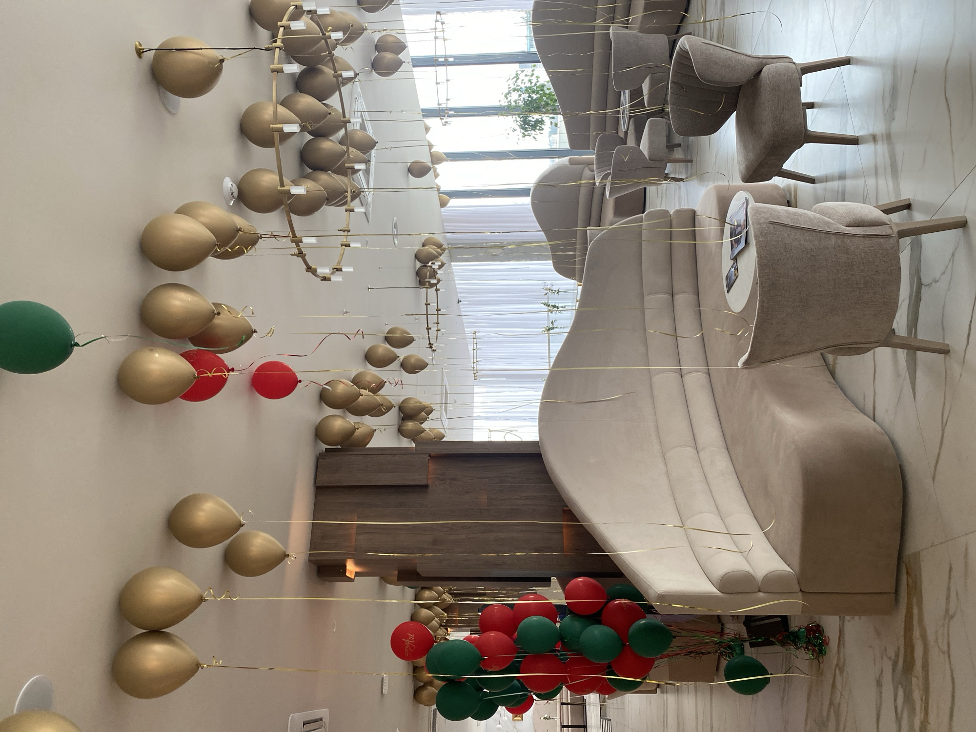 The image shows an interior space, possibly a lobby or lounge area, in Sochi, Russia. There is a long, curved beige sofa on the right, with chairs placed around it. The floor is a light-colored marble or tile. The wall on the left is decorated with gold balloons attached with clear rods, extending from the wall towards the center of the room. There are also clusters of red and green balloons nearer to the bottom of the image. Gold ribbons are attached to the balloons, trailing down towards the floor. There are large windows in the background, suggesting a view of the city or surrounding landscape. A wooden structure is visible between the sofa and wall decorations. The room appears unoccupied.