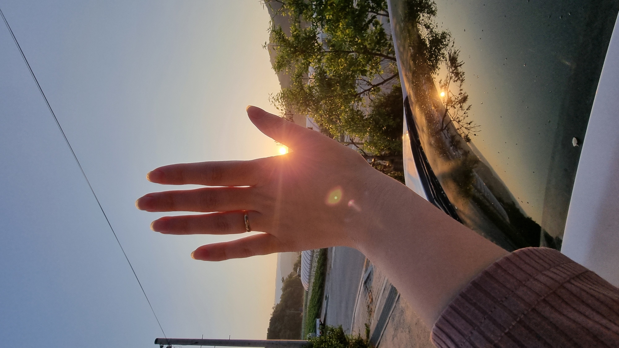 A low-angle shot of a hand held up to block the sun. The hand is light-skinned and has a silver ring on the ring finger. The sun is shining through the hand, creating a starburst effect between the fingers. In the background, there is a blue sky, trees, a road, and a car hood that reflects the trees and sunlight. A utility pole with wires is visible in the distance. The location is Dunnae-myeon, South Korea.