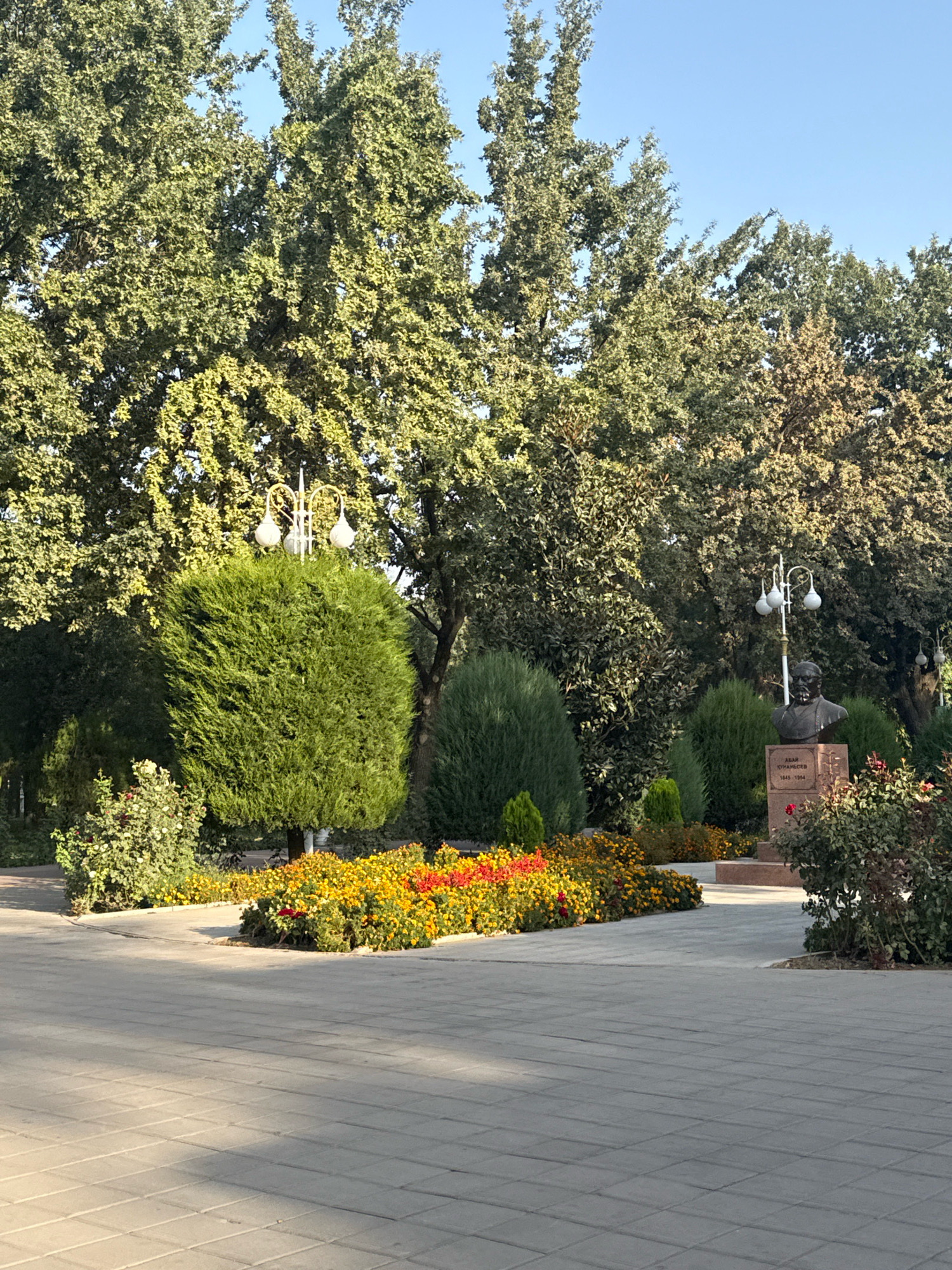 The image depicts a well-maintained park area in Dushanbe, Tajikistan, featuring a paved walkway in the foreground and a backdrop of lush greenery. A bust of Abai Qunanbaiuly, a prominent Kazakh poet and philosopher, is prominently displayed on a pedestal. Surrounding the bust are vibrant flowerbeds with a mix of yellow and red blossoms, and manicured evergreen shrubs. A decorative lamppost with three globe-shaped lights stands near a large, rounded evergreen tree. In the background, a row of mature trees with dense foliage forms a natural boundary, with a clear blue sky overhead. The scene is bathed in natural light, suggesting it was captured during daytime.