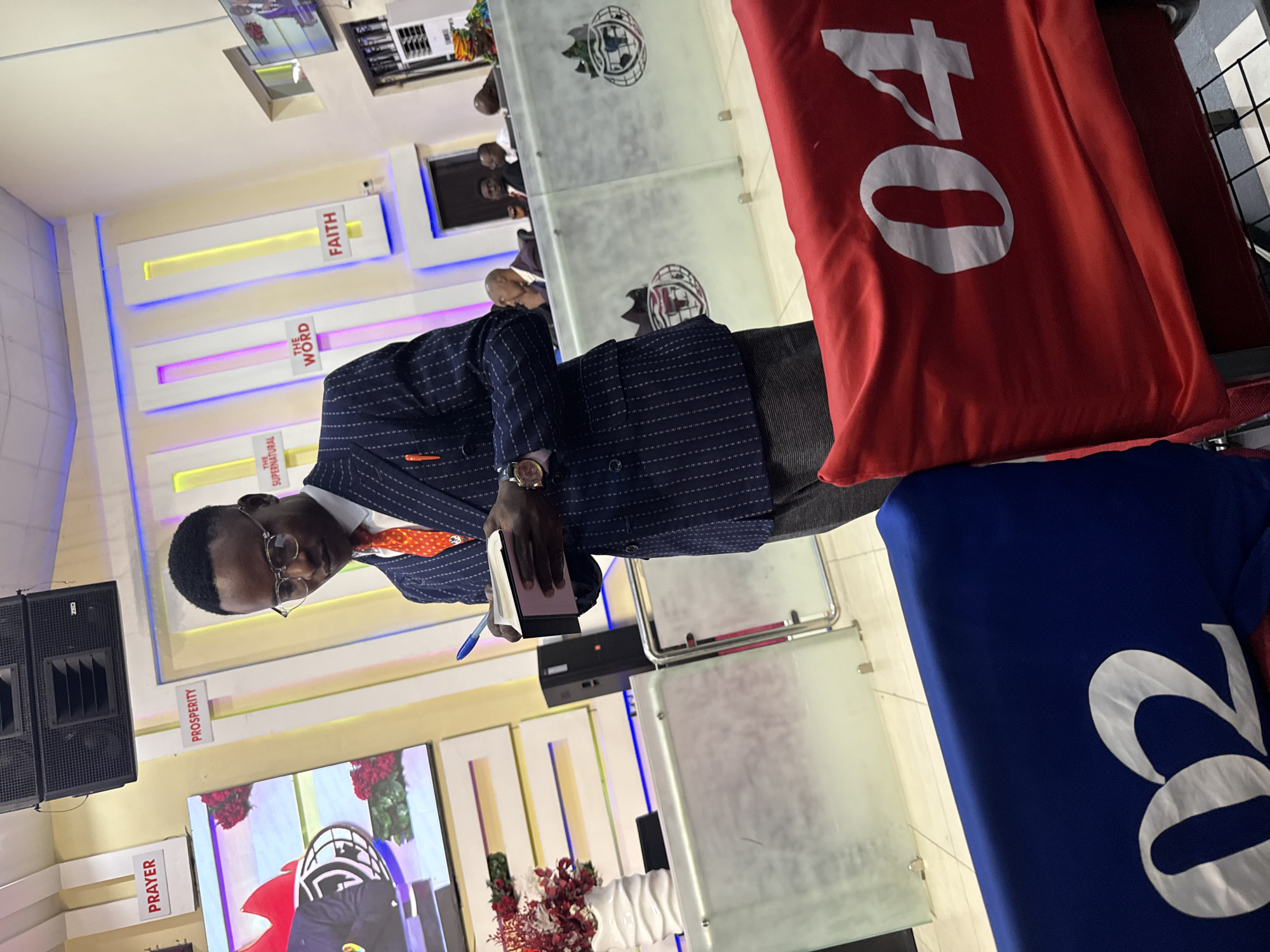 A man stands in an indoor setting, likely a church or religious venue in Lugbe, Nigeria. He is wearing glasses, a dark pinstripe suit with an orange tie and pocket square, and a wristwatch. He holds a notebook and pen, possibly taking notes. The environment features signage with words like "PRAYER," "PROSPERITY," "SUPERNATURAL," "WORD," and "FAITH" arranged vertically on the wall. In the background, there is a video screen showing what appears to be a religious broadcast. To the right of the man are items displaying the numbers "04" and "22". A transparent podium with a church logo is visible. Speakers stand nearby. Other individuals are visible in the blurred background.