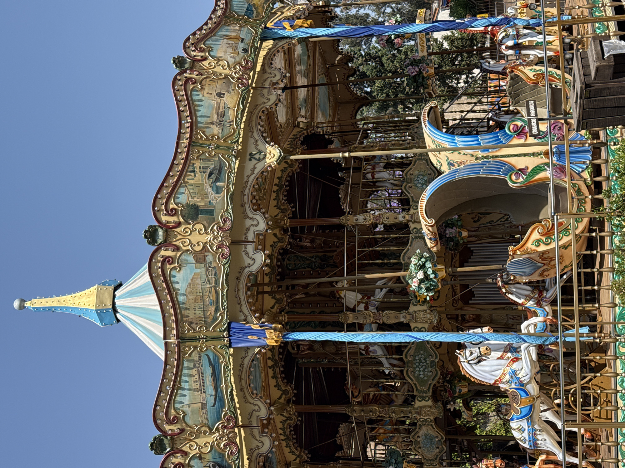 The image depicts a carousel, likely photographed in daylight due to the bright blue sky visible in the background. The carousel features ornate carvings and paintings, suggesting a vintage or antique style. Several horses are visible, along with other decorative elements such as painted panels with landscape scenes and decorative lights. Based on the location information, it is likely situated in Domingo Arenas, Mexico. There are also boats, trees, and flower decorations adorning the carousel. The carousel appears stationary. There are no people visible.