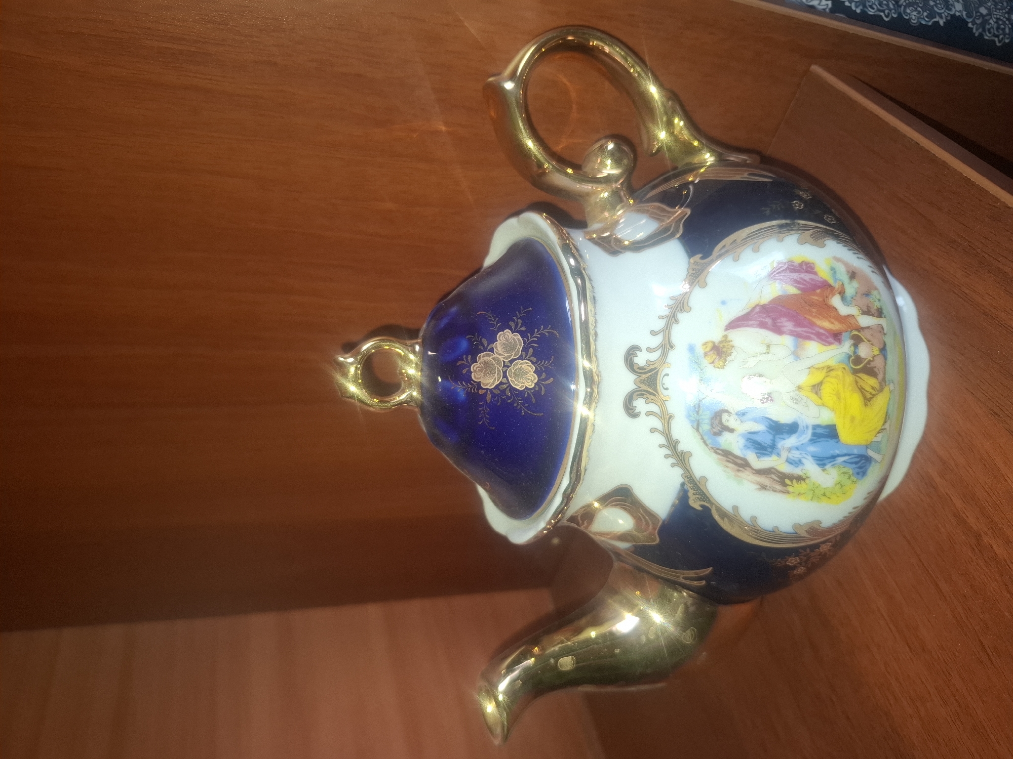 A vintage porcelain teapot with ornate gold accents and painted imagery sits on a wooden shelf. The teapot has a dark blue lid with gold floral designs, a white body with blue and gold decorative borders, and a gold spout and handle. The painted scene on the body depicts figures in classical attire. The background consists of a warm-toned wooden surface and shelving. The teapot appears to be displayed indoors, likely in a home or curio cabinet. There is no indication of the city of Almaty, Kazakhstan in the image.