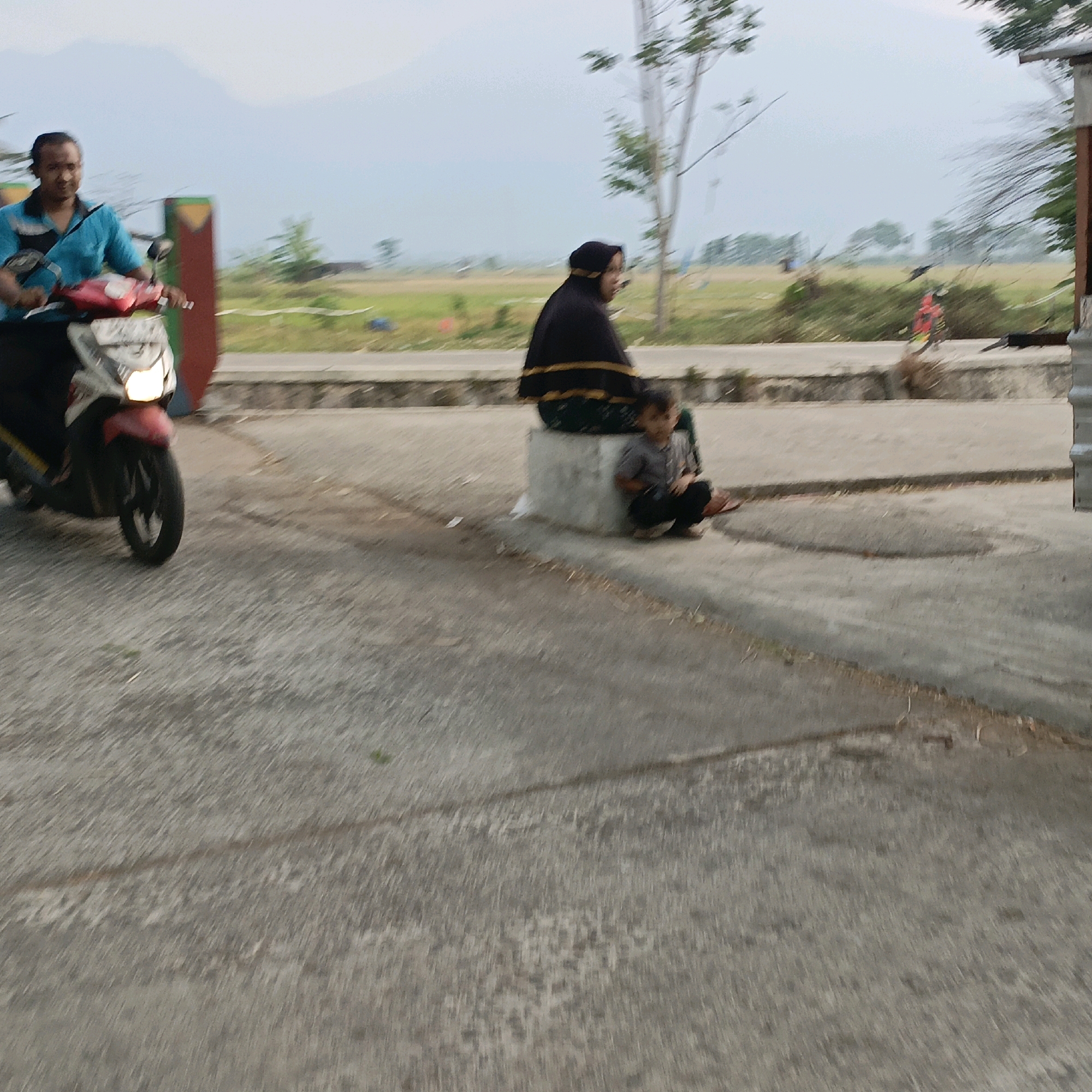 A person is riding a red and white motorcycle with its headlight on. The rider is wearing a blue shirt and black pants. Behind the motorcycle, a woman in a black hijab is seated on a concrete block. A child in a gray shirt and black pants is crouching next to the woman. The background shows a blurred landscape with a field and trees, suggesting a rural or semi-rural area. The image appears to be taken in Kesongo, Indonesia.
