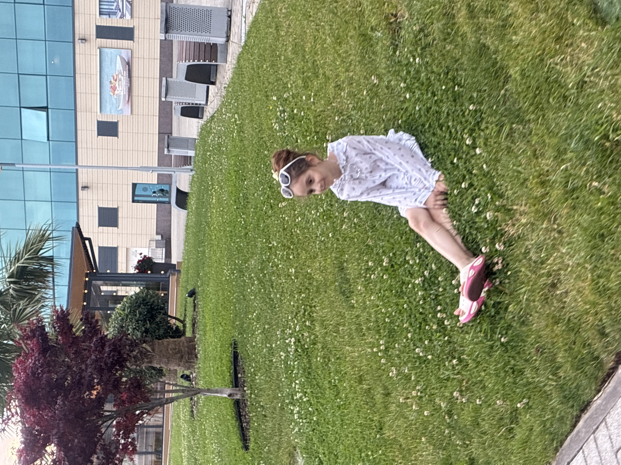 In Sochi, Russia, a young girl with her hair tied up in a bun and wearing white sunglasses on her head sits on a grassy lawn dotted with small white flowers. She is dressed in a white patterned dress and pink flip-flops. She is looking to her left, with a slight smile. In the background, a modern building with a glass facade stands behind a sidewalk with benches and trash receptacles. A tree with dark red foliage is visible to the left, and palm trees are present as well. The lighting suggests it is daytime.