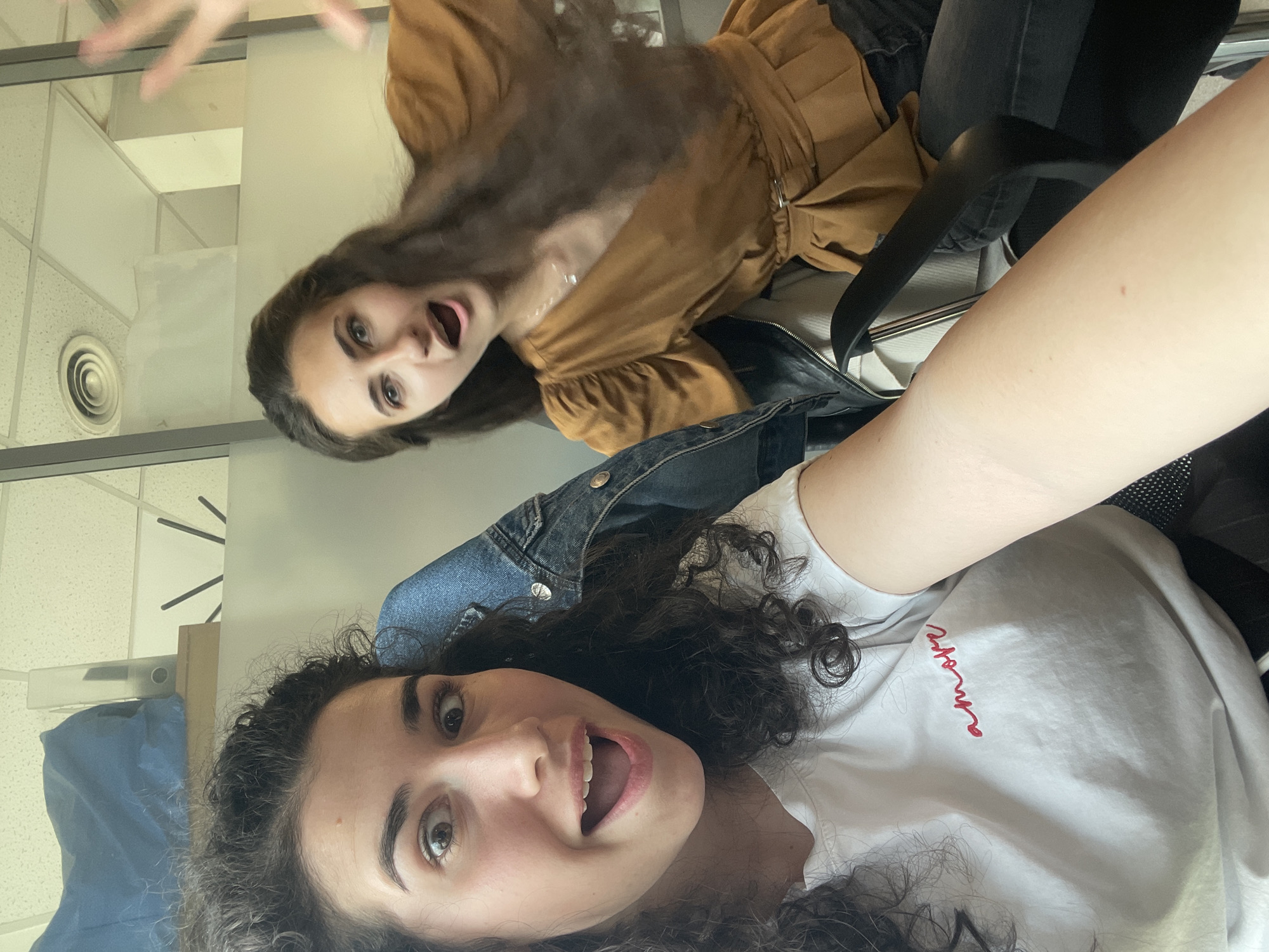 An upward-facing, inverted photo captures two young women indoors, likely in an office setting in Sofia, Bulgaria. The image is taken from a low angle, showing the women and the ceiling above them.
In the foreground, a woman with dark, voluminous curly hair looks directly at the camera with wide, light-colored eyes and an open mouth, conveying a playful or surprised expression. She wears a white t-shirt with indecipherable red cursive text on it and a blue denim jacket draped over her shoulders. Her bare arm is visible in the lower right of the frame.
Behind her, positioned further away and slightly to the left, another woman with similar dark, long hair is also looking at the camera with an open mouth and wide eyes, sharing a similar surprised or playful expression. She is wearing a brown, long-sleeved blouse with ruffled sleeves and a belted waist, paired with dark jeans. She appears to be sitting in a black office chair, which is partially visible.
The background shows typical office ceiling tiles, an air vent, and what appears to be a light fixture or shelf. Partially visible on the left side are a blue bag or object and abstract black lines that could be part of a wall clock. The overall mood is casual and lively.