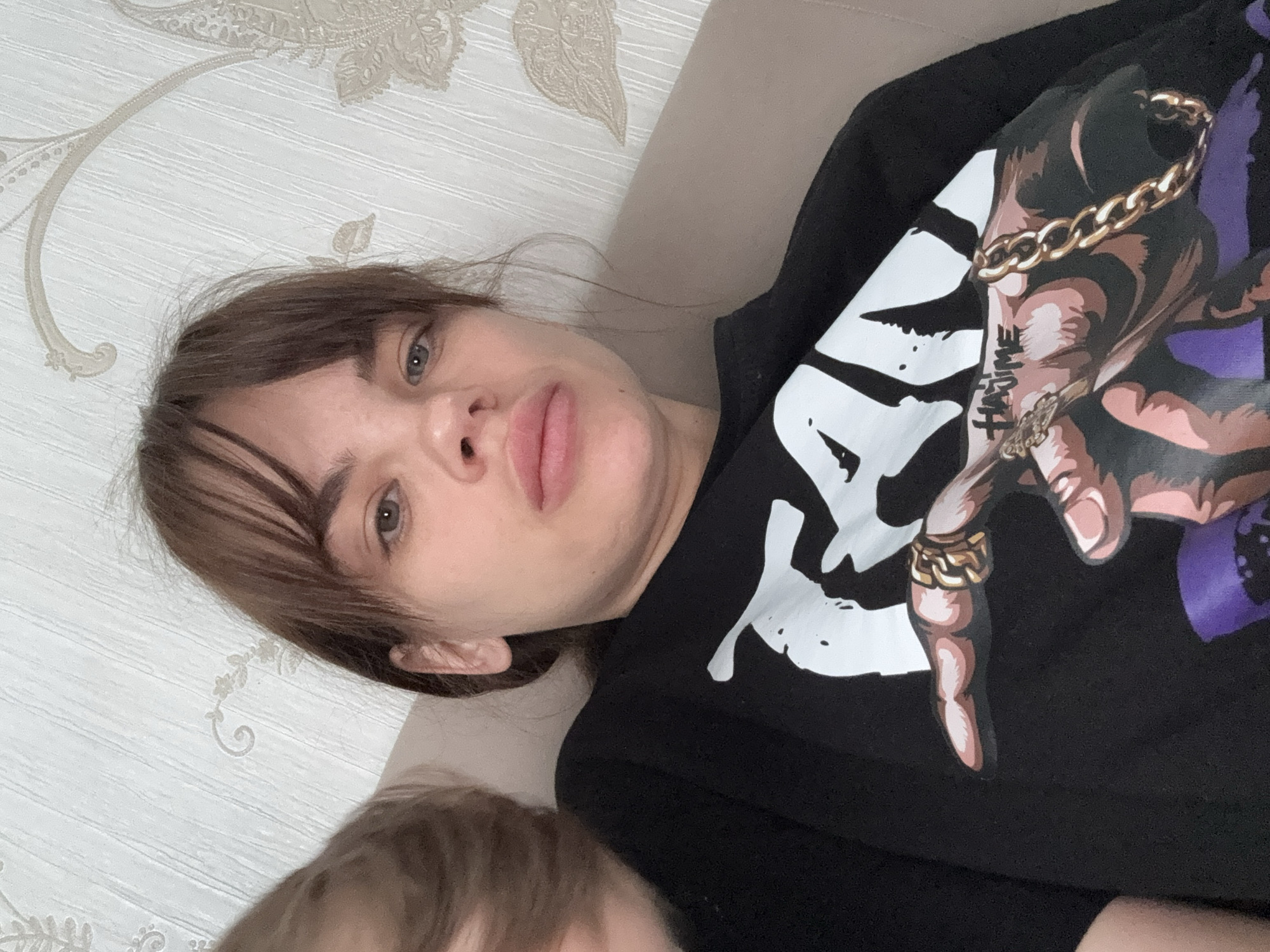 The image captures a close-up, angled shot of a woman lying down, looking directly at the camera, with a partial view of another person, likely a child, in the foreground. The woman has brown hair with bangs and a neutral to slightly pursed expression. She is wearing a black long-sleeved shirt with a prominent graphic design on the chest. The design features a stylized, cartoon-like hand, depicted in shades of brown and purple, wearing several gold rings on its fingers and a thick gold chain bracelet around its wrist. On the wrist of the graphic hand, there is visible text that reads "HYPE" in a stylized, black font.
The woman is positioned on a light beige or off-white upholstered surface, likely a couch or bed. Behind her is a wall covered with light-colored wallpaper that has a subtle, textured pattern of ornate floral or paisley motifs. The top of another person's head, with light brown hair, is visible in the lower left corner of the frame, suggesting the presence of a child close to the woman. The scene takes place indoors, likely within a home in Rostov-on-Don, Russia, portraying a casual, relaxed moment. The time of day is not discernible.