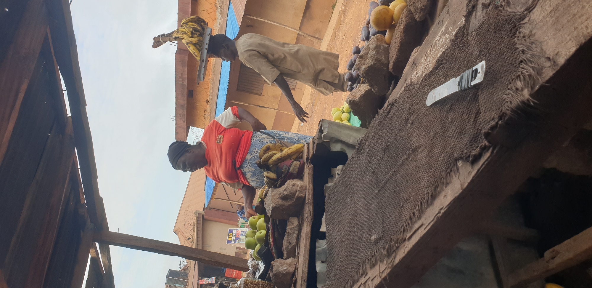 In the city of Yelwa, Nigeria, two individuals are depicted at an outdoor market stall. An overweight woman, wearing an orange t-shirt and a blue patterned skirt, stands behind a wooden counter laden with produce. To her right, a slender man, dressed in a light brown tunic, walks away from the stall, balancing a tray of bananas on his head. The stall itself is constructed from rough wooden planks and covered by a corrugated metal roof, supported by wooden beams. In the foreground, a rough burlap cloth covers part of the wooden counter, and a knife with a silver blade and wrapped handle rests on it. Behind the stall, a brown building with a blue roof and barbed wire fencing is visible, along with other market stalls and a water tank. The ground is covered in reddish-brown dirt, characteristic of the Nigerian landscape. The sky above is overcast with a few scattered clouds.