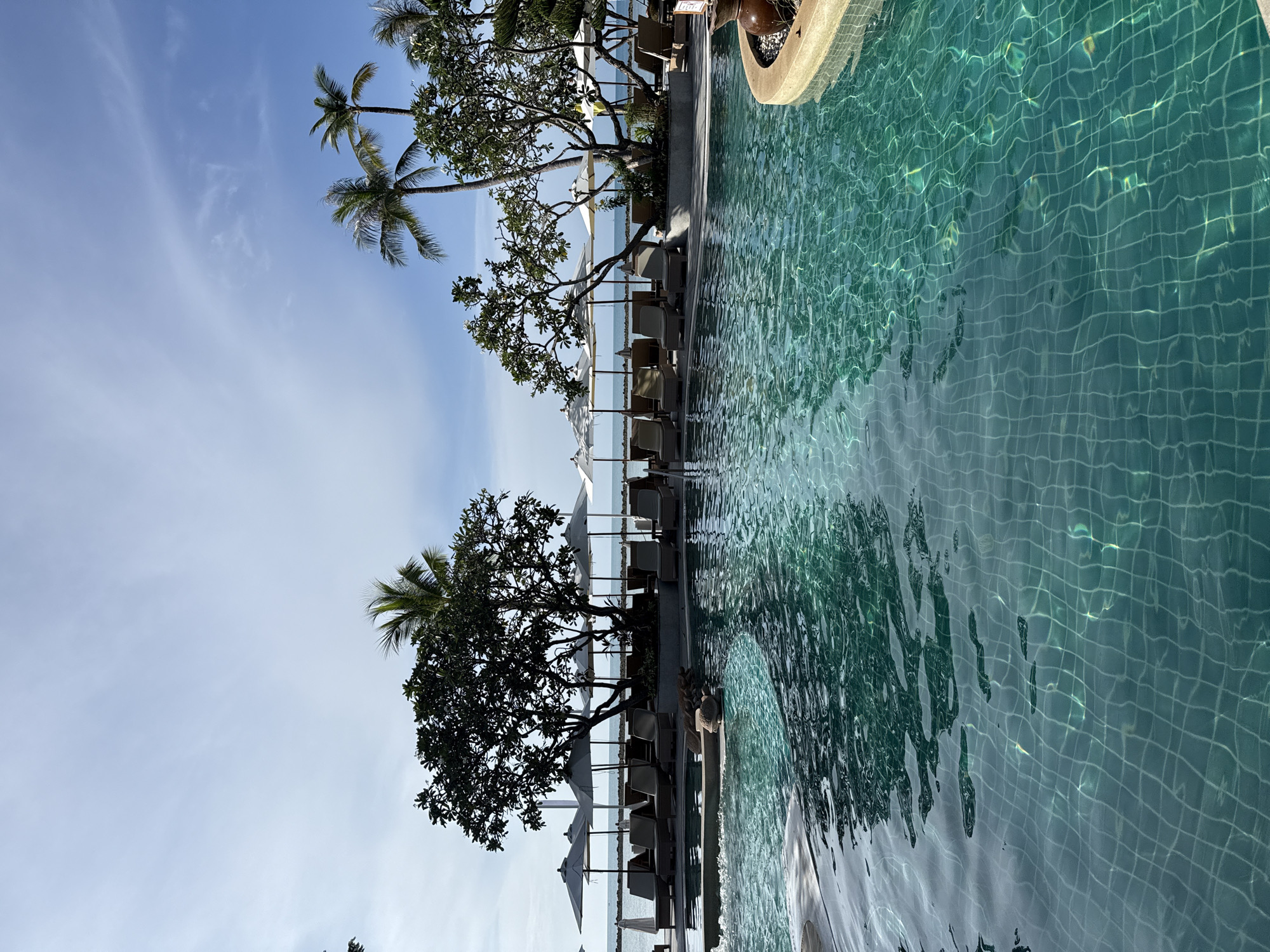 A resort swimming pool is in the foreground of the image, with the turquoise water reflecting sunlight. In the background, there are lounge chairs, umbrellas, and palm trees. The ocean is visible beyond the pool area, with a clear sky overhead. The resort appears to be located on the island of Ko Samui, Thailand, likely in the Chaweng or Lamai beach area. There are no people visible in the immediate foreground.