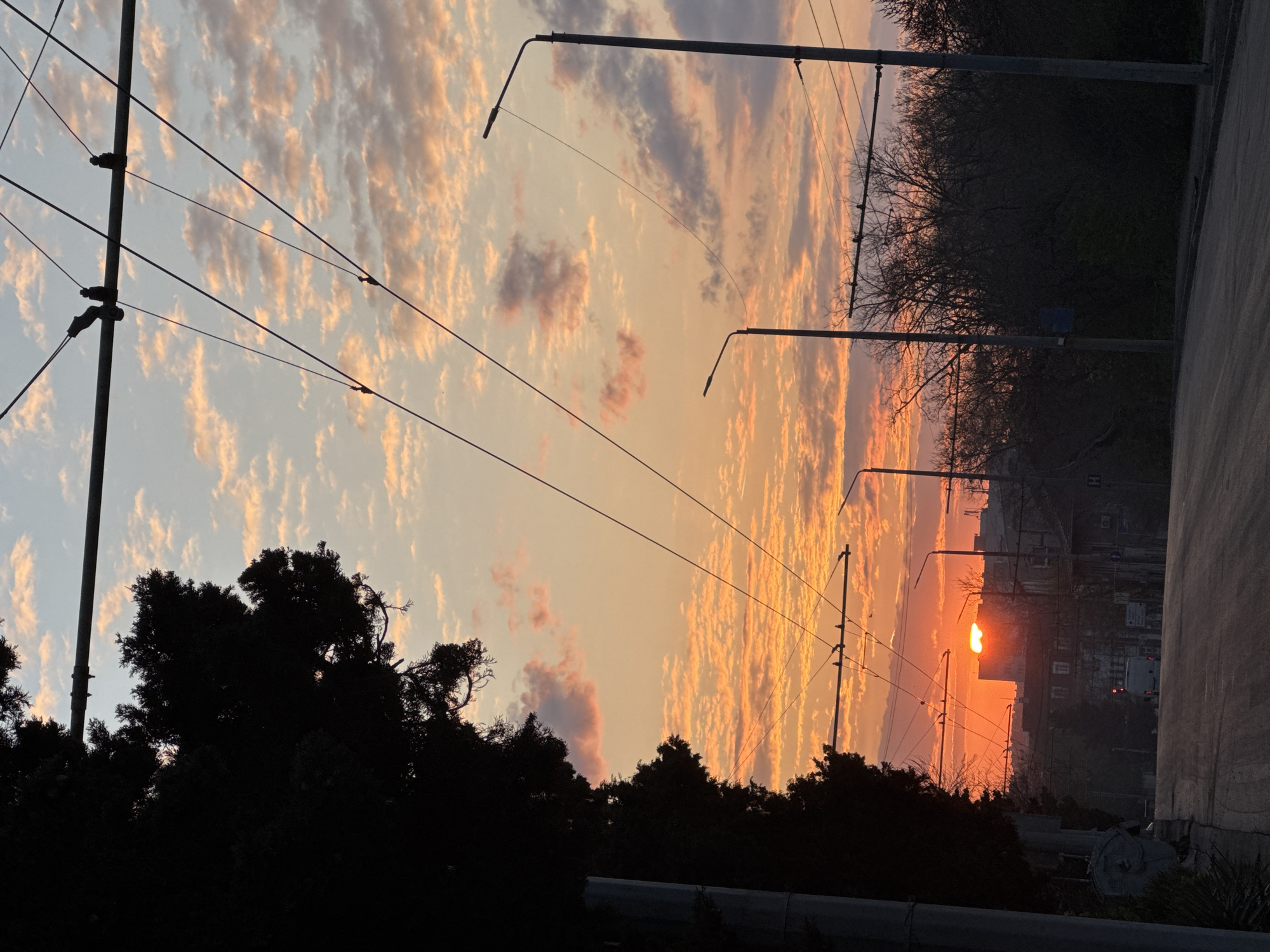 The image captures a sunset in Burgas, Bulgaria, with a low sun appearing between buildings. Foreground elements include silhouetted trees and street lamps against the vibrant orange and pink sky. Overhead power lines extend across the top portion. The right side shows a paved surface suggesting a road or street. The sky features scattered, textured clouds illuminated by the setting sun.