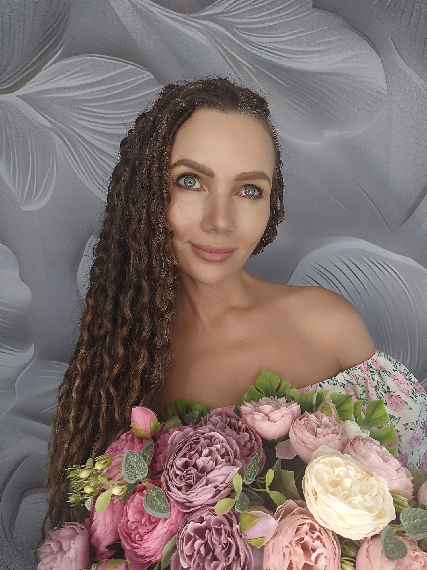 A woman with long, curly brown hair is posing for a portrait, holding a bouquet of pink and cream flowers. She is wearing an off-the-shoulder floral top. The background is a textured grey wall with a pattern of abstract, leaf-like shapes in a lighter shade of grey. Her eyes are a striking blue, and she has a gentle smile. The lighting appears to be soft and even, suggesting an indoor setting. The overall mood is serene and feminine, emphasizing the beauty of the flowers and the subject. There is no visible text in the image.