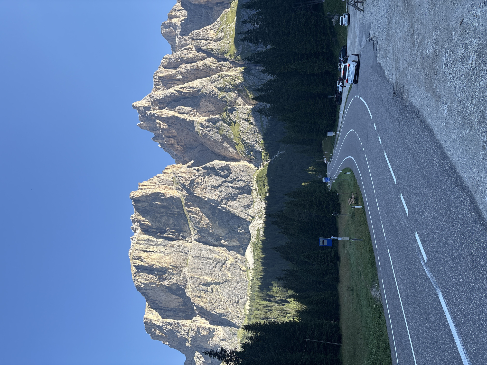 This is a photograph of the Sella Pass in Canazei, Italy, taken on a clear day under a bright blue sky. The foreground features a paved, winding road with white lane markings. To the right of the road, a gravel parking area is visible with several parked cars, including a white SUV. Beyond the road and parking area, a dense forest of dark green pine trees stretches across the midground, their tops forming a dark line against the lighter green lower slopes of the mountains. The background is dominated by a massive, rugged mountain range, characterized by sheer rock faces, sharp peaks, and dramatic shadows. The mountains are bathed in sunlight, highlighting their textured surfaces and the sparse vegetation clinging to their lower sections. In the distance, the sky is a clear, uniform blue.