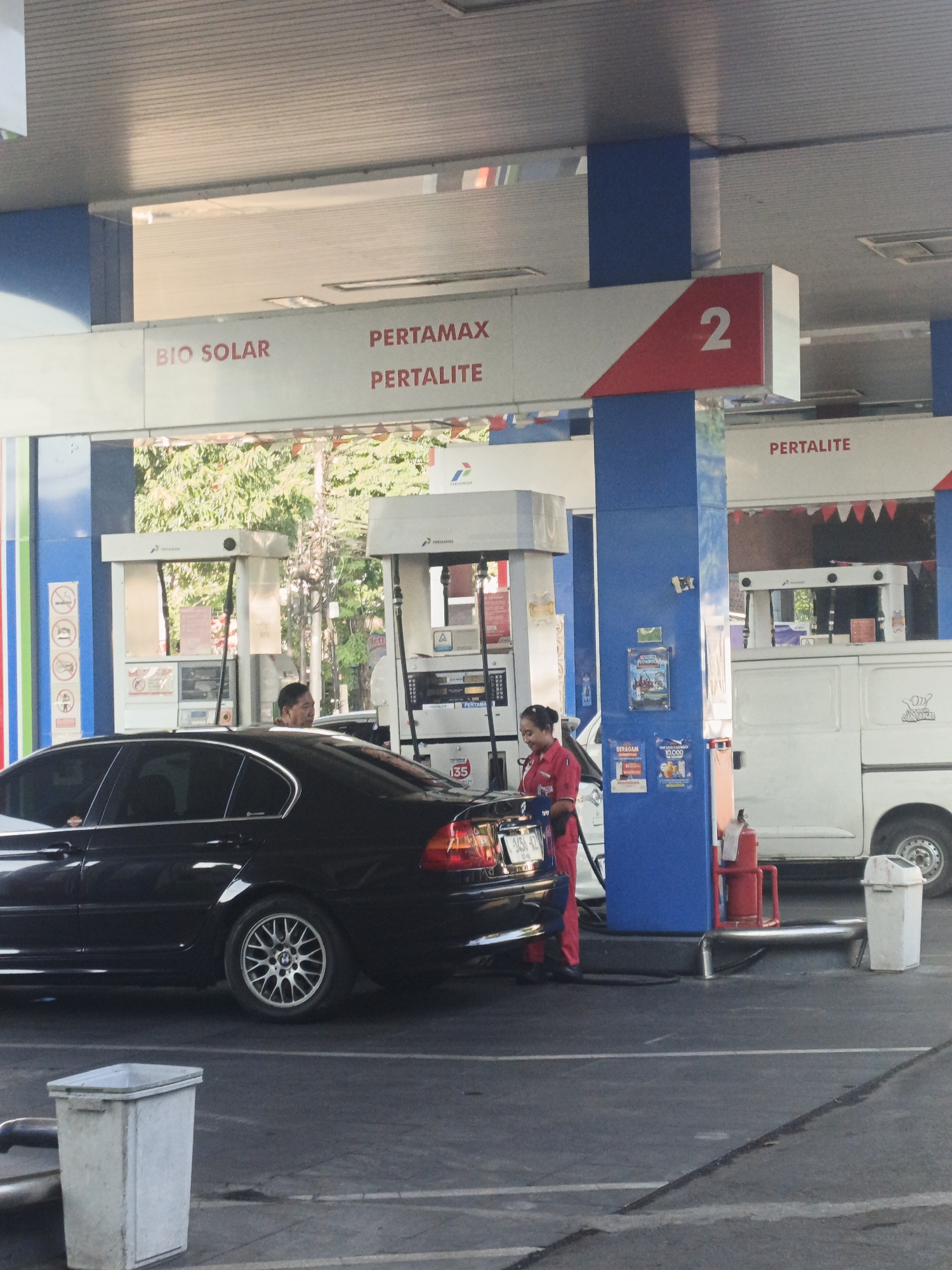 This is a medium shot of a gas station in Surabaya, Indonesia, with a black car being refueled. A female attendant in a red uniform is operating the gas pump. To the left of the car, a white trash can is visible. Behind the car and the pump, a white service vehicle is parked. The gas station features blue and white signage with red accents, indicating fuel types and a pump number "2". Trees and foliage are visible in the background.