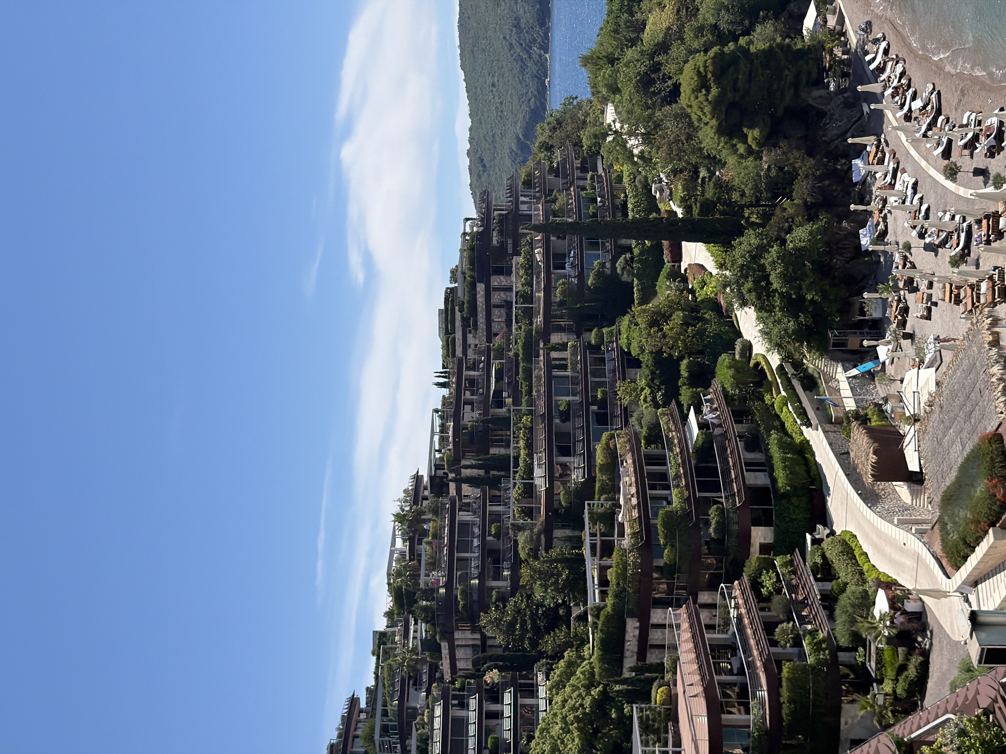 The image captures a resort complex built into a hillside overlooking a bay in Budva, Montenegro. The architecture features multiple levels of terraces, each adorned with lush greenery and private balconies. The buildings are a mix of modern design with wooden accents and stone facades, blending harmoniously with the natural landscape.
The foreground shows a beach area with rows of lounge chairs and umbrellas, some occupied by sunbathers. A winding pathway leads from the beach up the hillside towards the resort. The bay is calm and a lush green mountainous coastline is visible in the distance. The sky is a clear blue with a few wispy clouds. The scene appears to be set on a sunny day, suggesting a vacation or leisure setting.