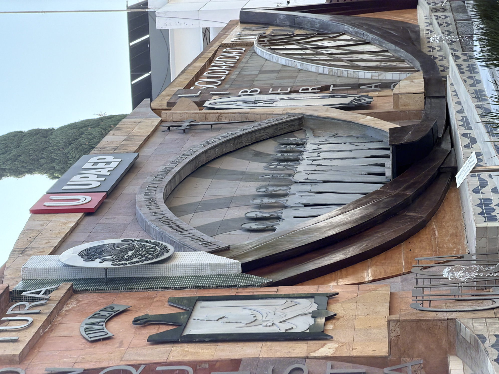 The image captures a portion of the UPAEP (Universidad Popular Autónoma del Estado de Puebla) monument facade in Puebla, Mexico. The facade consists of layered, dimensional installations with text, symbols, and figures. Words "UPAEP," "SOLIDARIDAD" "LIBERTAD" are visible. A sculptural depiction of a row of figures stands in the center. A large, circular element is above them. Below the figures are two additional sculptural installations. One is a circular relief of the Mexican coat of arms (eagle devouring a serpent), and the other appears to be an allegory. The monument is constructed from stone, metal, and tile. The facade is situated against a backdrop of buildings and greenery.