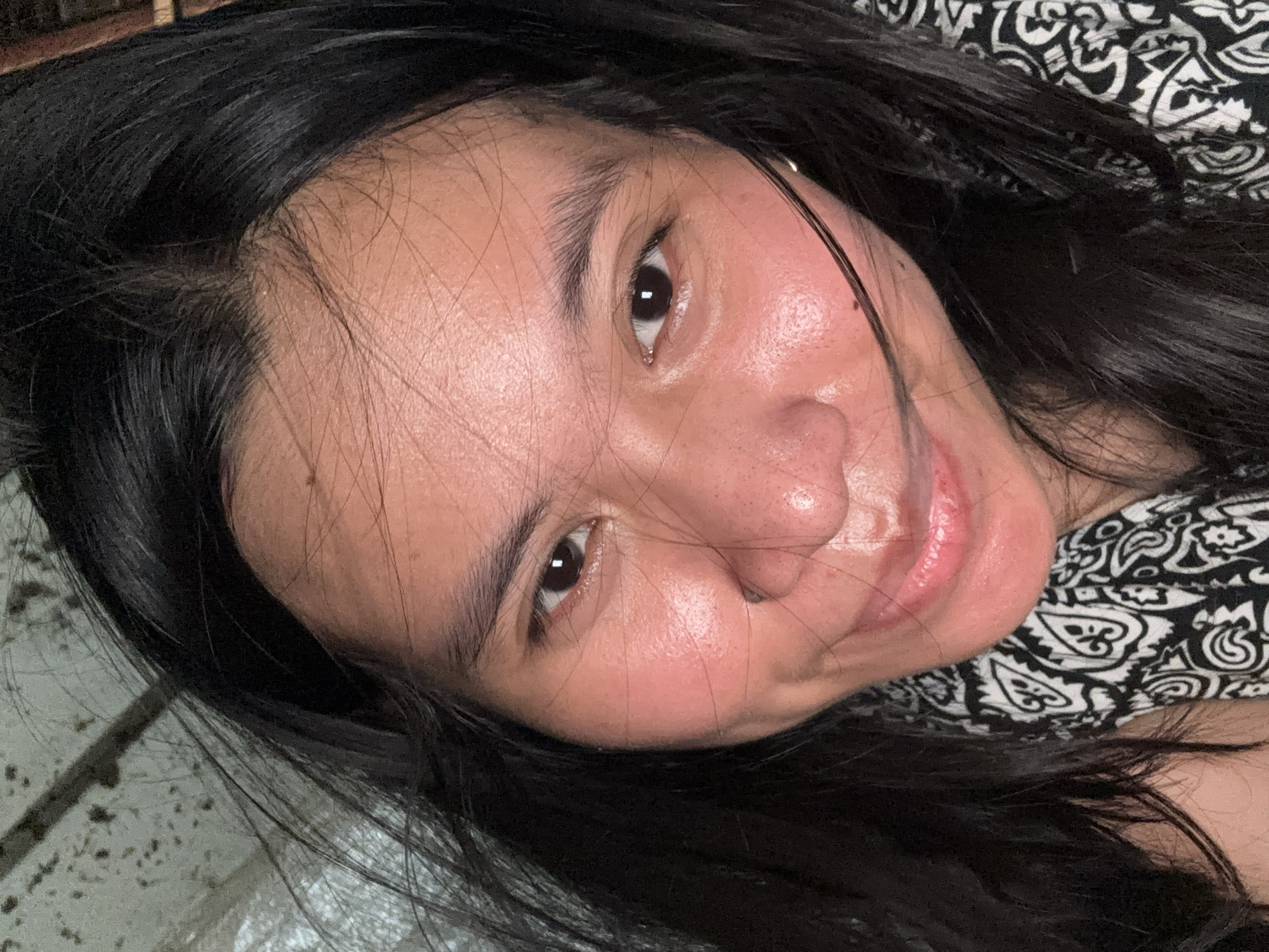 A close-up, overhead selfie shows a young woman with long, dark hair. She is wearing a black and white patterned garment around her neck. The background appears to be a simple wall or ceiling. The image was taken in East Jakarta, Indonesia.