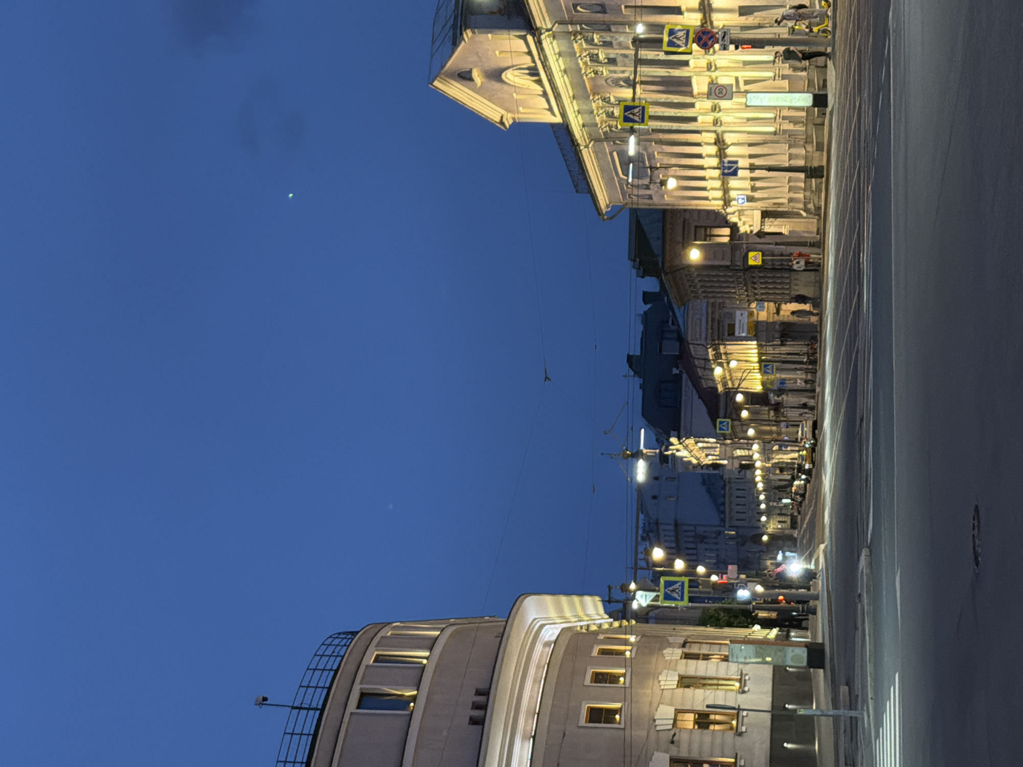 The image captures a street view in Moscow, Russia, at dusk or night. On either side of the road, buildings illuminated with artificial light contrast against the darkening blue sky. Pedestrian crossings are marked with signs. People are visible, though details are obscured by the lighting. The road surface reflects light, indicating it may be wet.