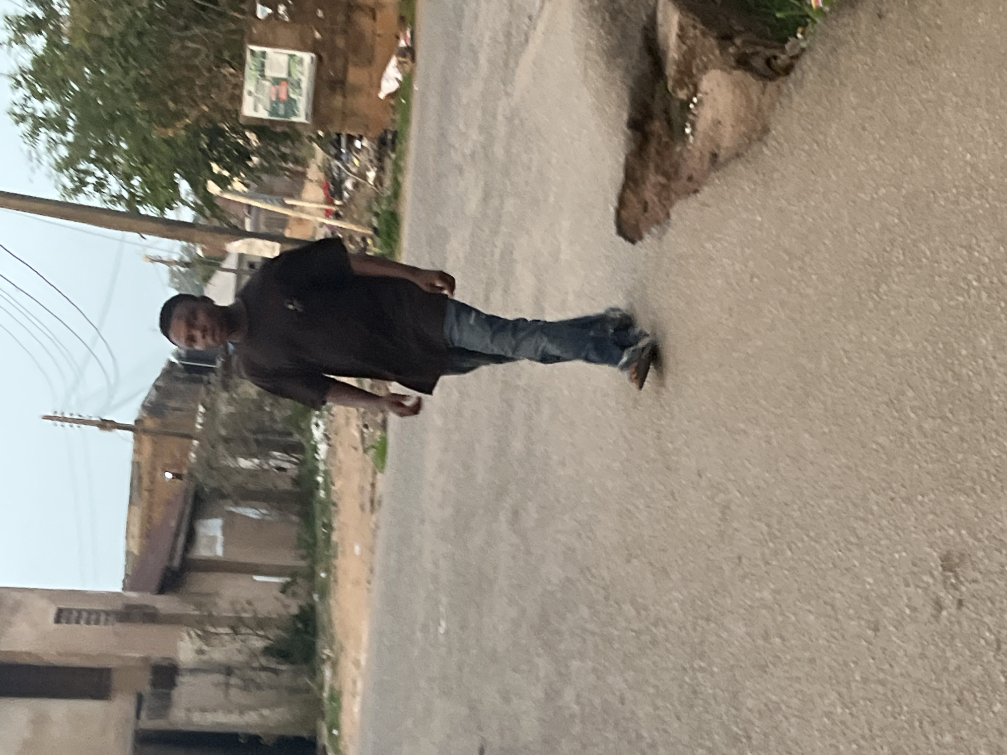 A person in a dark shirt and blue jeans is walking along a paved road in Jos, Nigeria. To the left, there are buildings with visible windows and a complex of electrical wires. On the right, the road has an elevated edge with some vegetation visible. In the background, there is a sign and general urban debris, suggesting an urban or semi-urban environment. The overall tone is neutral and documentary.