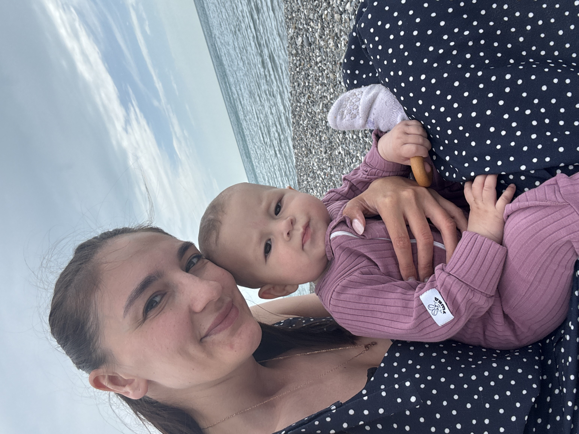 The image shows a woman holding a baby in her arms. The woman is smiling and looking at the camera. The baby is looking at the camera with a neutral expression. The woman is wearing a dark blue dress with white polka dots and a gold necklace. The baby is wearing a purple ribbed outfit. The baby is also wearing a white sock. In the background, there is a pebble beach, the sea, and a cloudy sky. The photograph appears to be taken in Sochi, Russia.