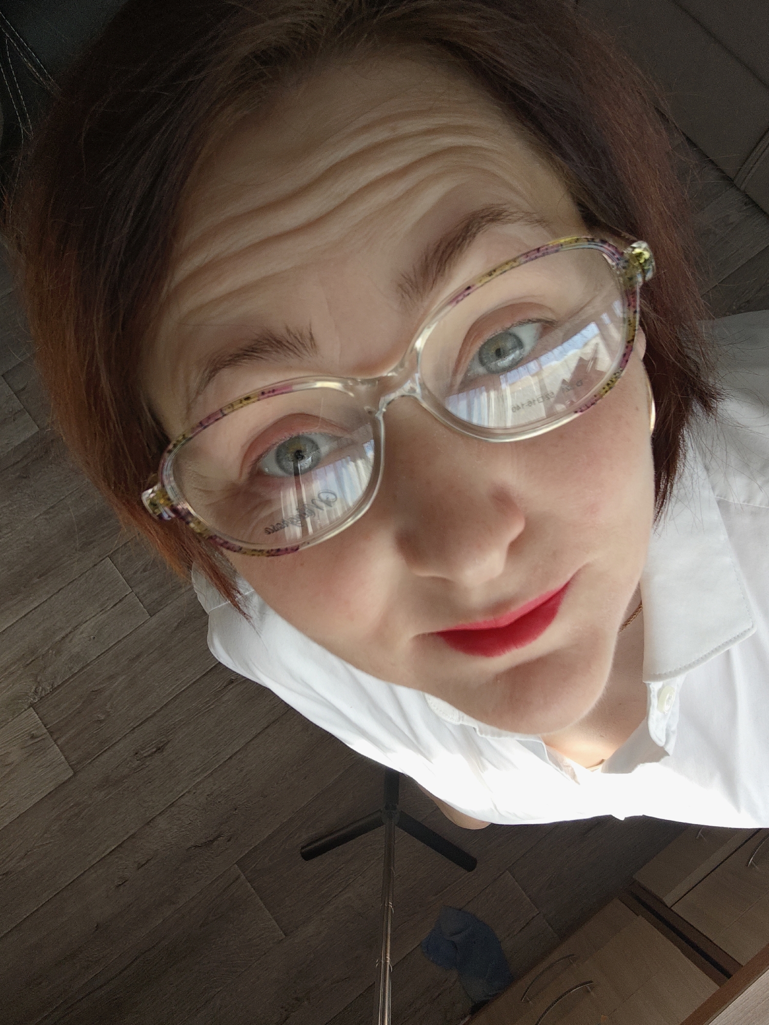A woman with auburn hair and fair skin is centered in the frame, looking directly at the camera from a low angle. She is wearing glasses with decorative frames. Her lips are painted bright red. She is wearing a white collared shirt. The background consists of a wooden floor and a small portion of a cabinet. It is not possible to determine if this image was taken in the city of Markovskii, Russia from the contents of this photo.