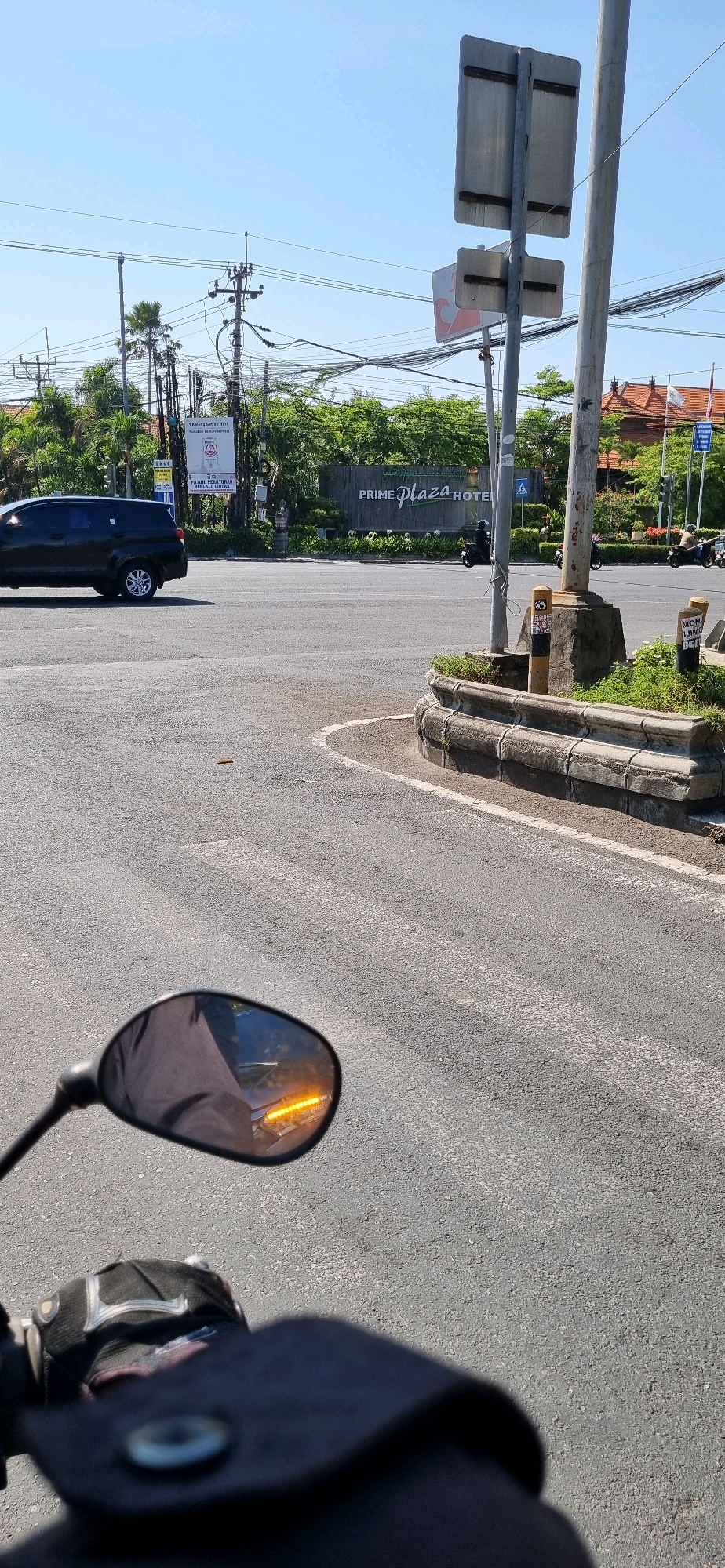The image captures a street scene in Denpasar, Indonesia, likely at an intersection, dominated by a bright, sunny sky and asphalt road. In the foreground, the perspective is from a motorcycle, with the handlebars and a portion of the rider's glove and jacket visible. A side-view mirror reflects an orange turn signal light, and the road surface behind the motorcycle.
Further back, a black SUV is visible on the left side of the road. Across the intersection, a large sign displaying "PRIME PLAZA HOTEL" is prominent, surrounded by lush green trees. A traffic light is visible on the right side of the image, and several motorcycles are in motion on the road, indicating ongoing traffic. Utility poles with numerous wires crisscross the upper portion of the image, typical of urban infrastructure. The architecture of buildings with red-tiled roofs can be seen behind the trees on the right. The scene appears to be during daylight hours, with strong shadows cast on the road.