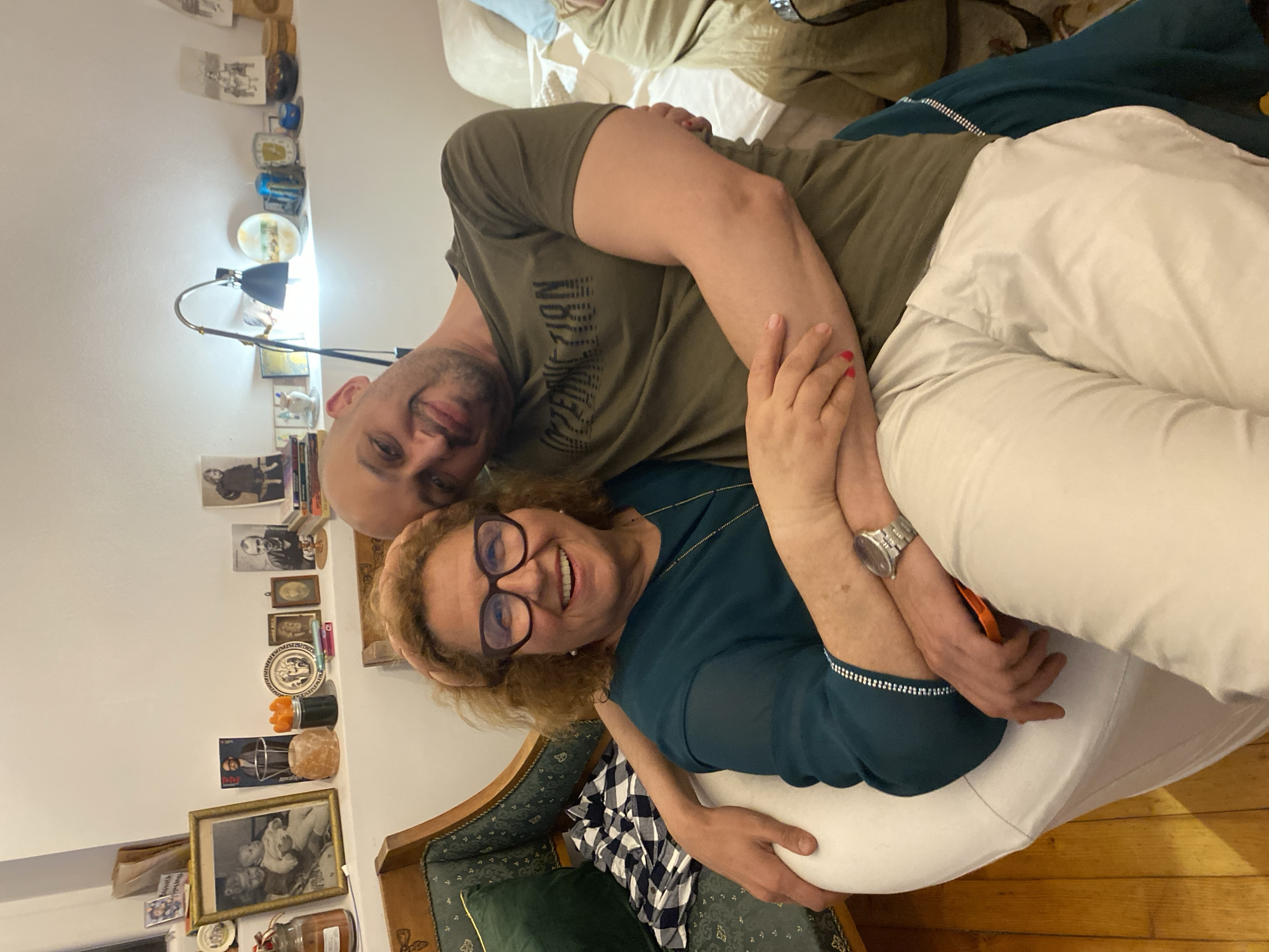 Two people are posed together indoors. A man with a shaved head is wearing a green t-shirt. A woman with curly hair and glasses is wearing a teal top and white pants. They are embracing. A lamp is visible in the background, as are various decorative items on a wall. A patterned chair is also visible. The location appears to be a home interior. There is no indication in the image or context that it is in the city of Sofia, Bulgaria.