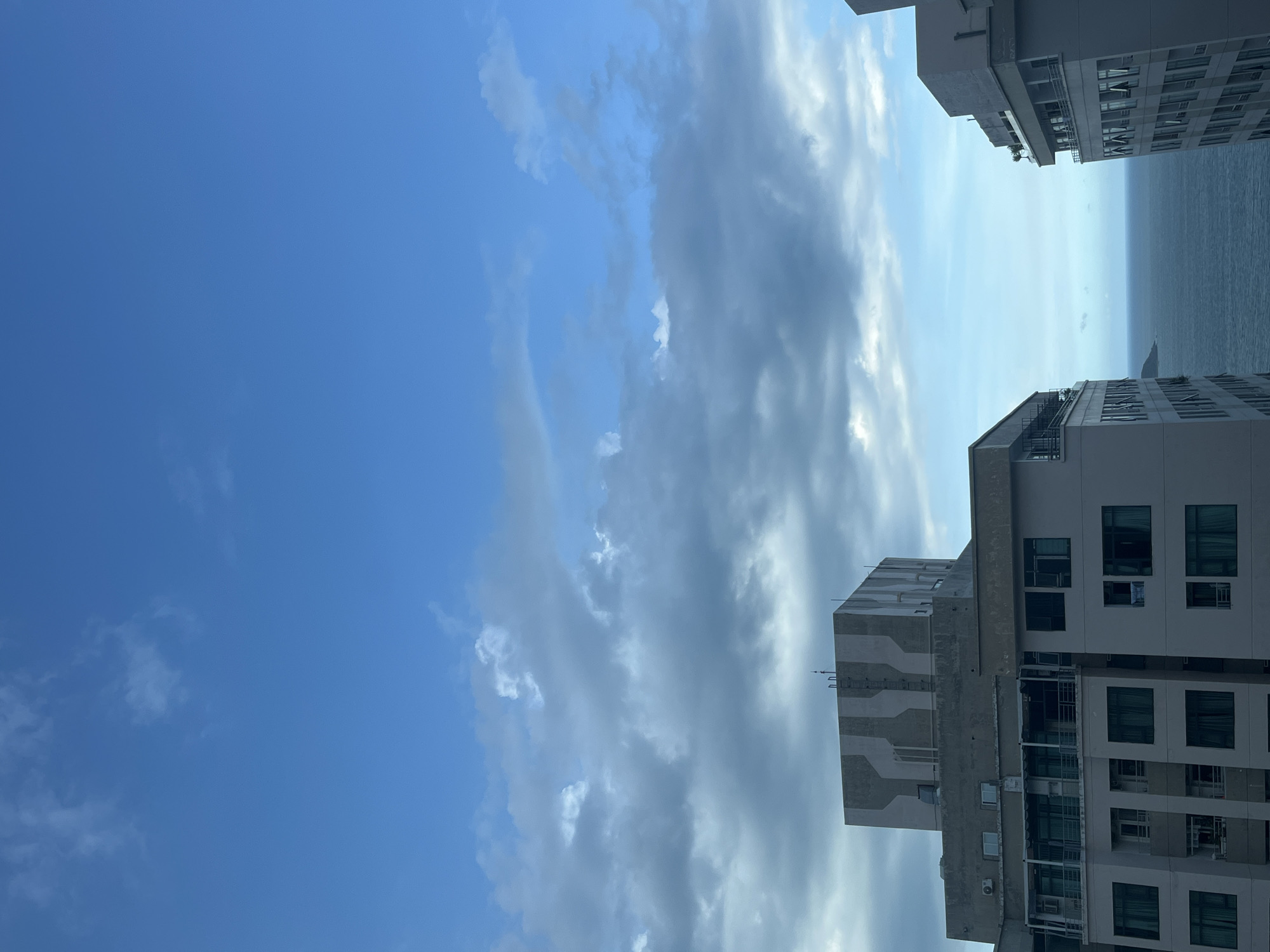 The image shows a view from what appears to be a high-rise building in Bac Nha Trang Ward, Vietnam. The scene includes a bright blue sky with patches of clouds, tall buildings, and a glimpse of the ocean. The buildings are modern, with light-colored facades and multiple windows. The ocean appears calm with light surface texture. The sky is predominately clear blue on the left side of the frame.