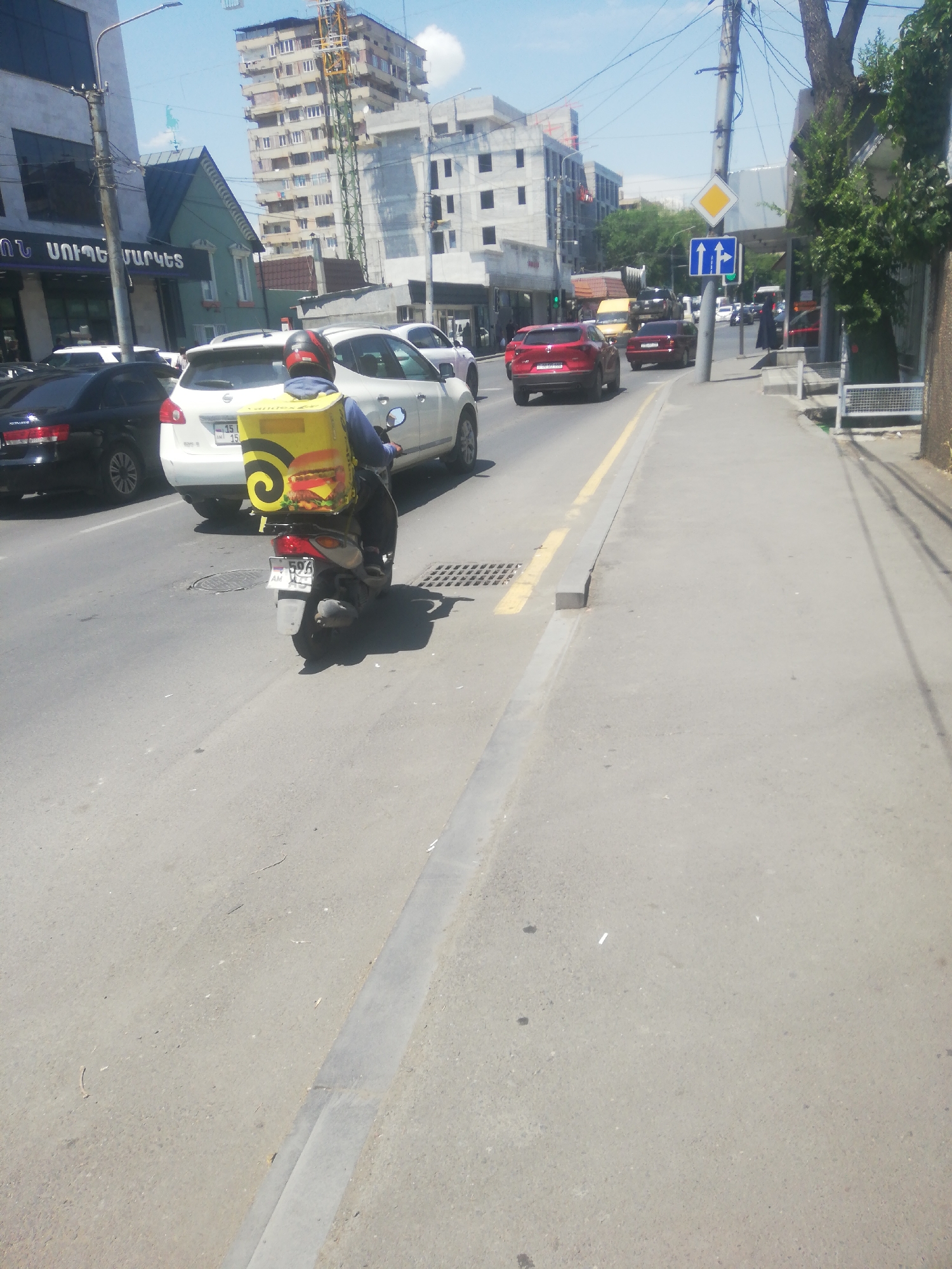 The image depicts a street scene in Yerevan, Armenia. In the foreground, a person wearing a red helmet rides a scooter with a large yellow delivery box on the back, which features an image of a hamburger. The scooter has a license plate that is partially visible. The scooter is on the right side of a two-lane road. Several cars are visible on the road, including a white SUV, a black sedan, and red cars. In the background, buildings under construction and older residential buildings are present, along with trees and streetlights. Traffic signs are also visible. The sky is clear and blue, indicating a sunny day.