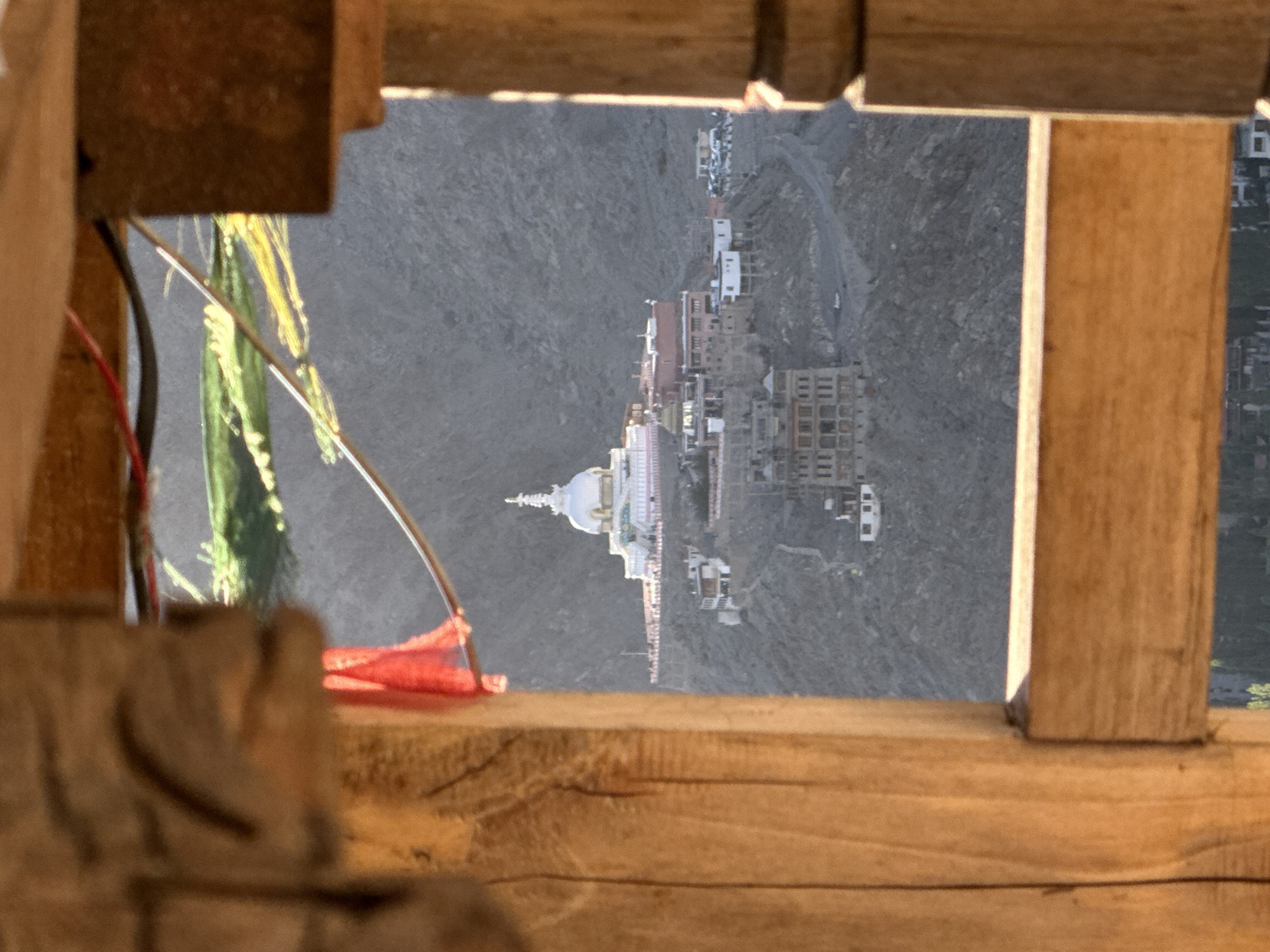 The image, captured in Leh, India, shows a view of the Shanti Stupa framed by wooden structures. The Shanti Stupa, a white domed Buddhist monument, is situated on a hill in the distance, surrounded by a complex of buildings and pathways. The foreground features weathered wooden beams, with a sliver of red fabric visible on the left. Prayer flags, adorned with yellow and green colors, are strung across the upper part of the frame. The background consists of a barren, rocky mountainside. The overall scene is bathed in the warm light of what appears to be either sunrise or sunset, casting long shadows and highlighting the texture of the wooden elements. There are no discernible people in the image.