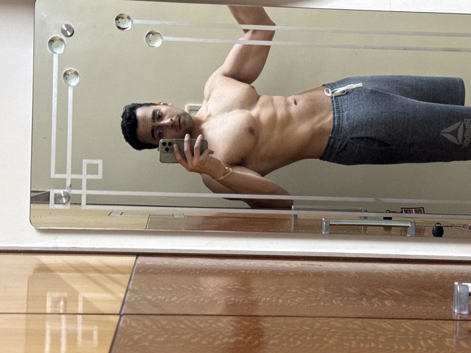 A man poses shirtless in a mirror selfie. He wears gray Reebok sweatpants. His physique is muscular with visible abs. He holds a smartphone in his left hand, his face in the reflection is visible. The mirror has a decorative etched pattern with circular accents. The background is a beige wall, and a polished wooden surface is below the mirror.