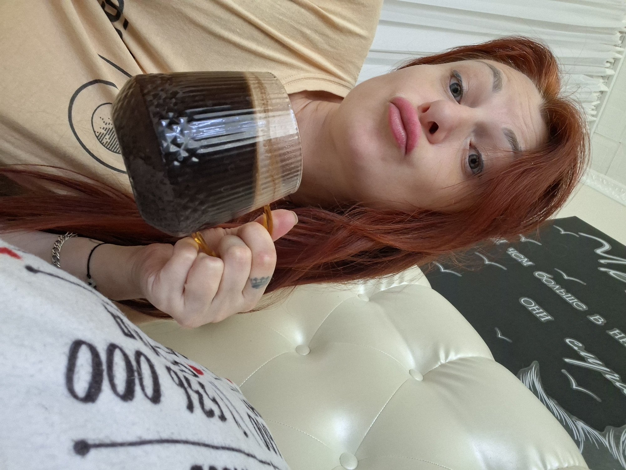 A medium shot captures a woman with auburn hair and rosy lips, making a pouty expression while holding a dark beverage in a ribbed glass mug. She is wearing a light brown t-shirt with a black graphic design. Her left hand, adorned with a ring, holds the mug, while her right hand rests near her chin. A tattoo is visible on her left hand. She is positioned in front of a white tufted headboard and a wall with black decorative lettering and bird silhouettes. The environment suggests an indoor setting, possibly a bedroom, with white curtains visible in the background. The image appears to be a selfie taken from a slightly low angle. The city of Myadzyel, Belarus, is not depicted in this image, nor is there any discernible information within the image that would place it in that specific location.