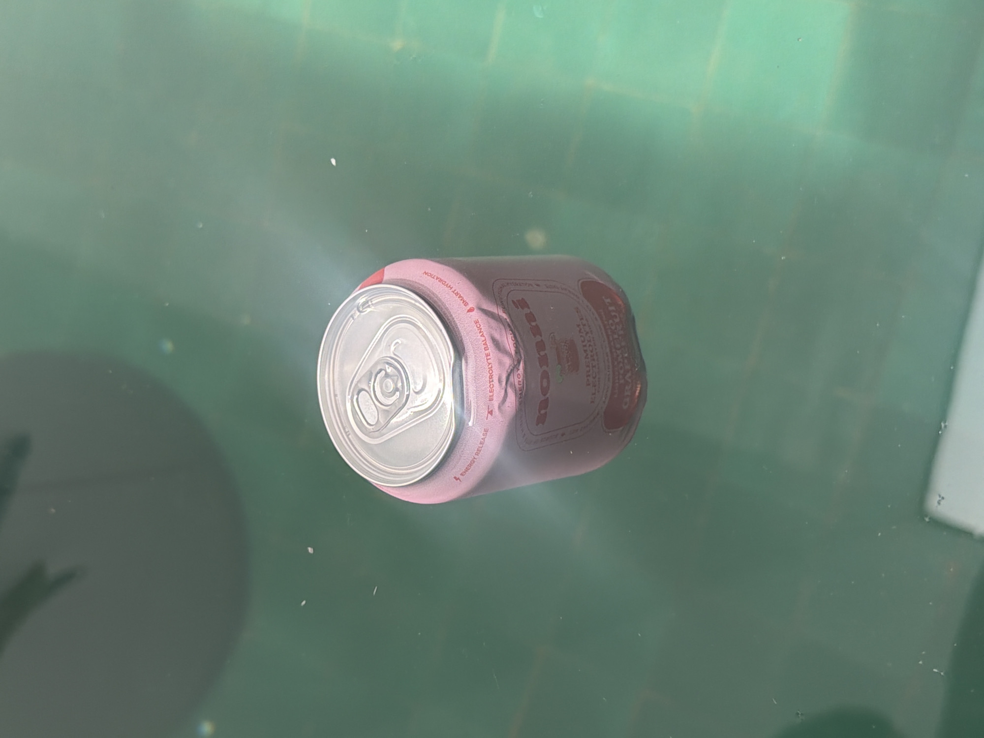 A pink beverage can with the brand name "nonni" is submerged in a greenish body of water, likely a swimming pool. The can is slightly dented and tilted, with its top opening facing upwards. The lid of the can is visible, showing the standard pull-tab mechanism. Text on the can indicates "ENERGY RELEASE," "ELECTROLYTE BALANCE," and "SMART HYDRATION," along with "PREMIUM ELECTROLYTES" and "POMELO GRAPEFRUIT." The water is clear, and there are some small white specks floating on the surface and suspended in the water. Shadows are cast on the surface of the water, suggesting overhead lighting or an object above the water. The overall environment appears to be an outdoor setting. There are no people visible in the image, and no specific landmarks or architectural features definitively place the scene in Marrakesh, Morocco.