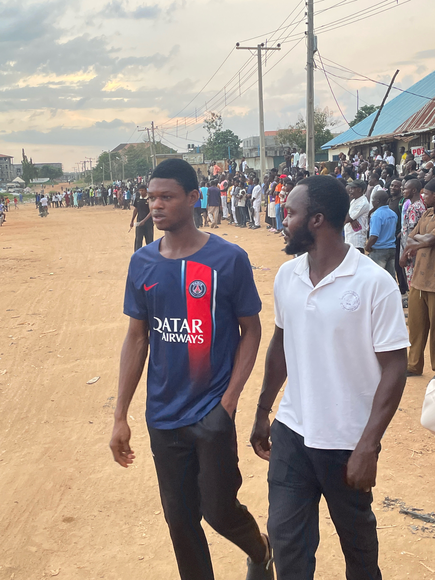 Two young men are walking on a dirt road in a crowded outdoor market or gathering in Abuja, Nigeria. The man on the left is wearing a dark blue and red soccer jersey with "QATAR AIRWAYS" visible on it, and black pants. The man on the right is wearing a white polo shirt with a circular logo on the left chest and dark pinstripe pants. Both men are walking towards the right of the frame. Behind them, a large crowd of people, predominantly men, are gathered, some standing and watching. The background shows a mixture of low-rise buildings, power lines, and a cloudy sky. The environment appears to be dusty and unpaved.