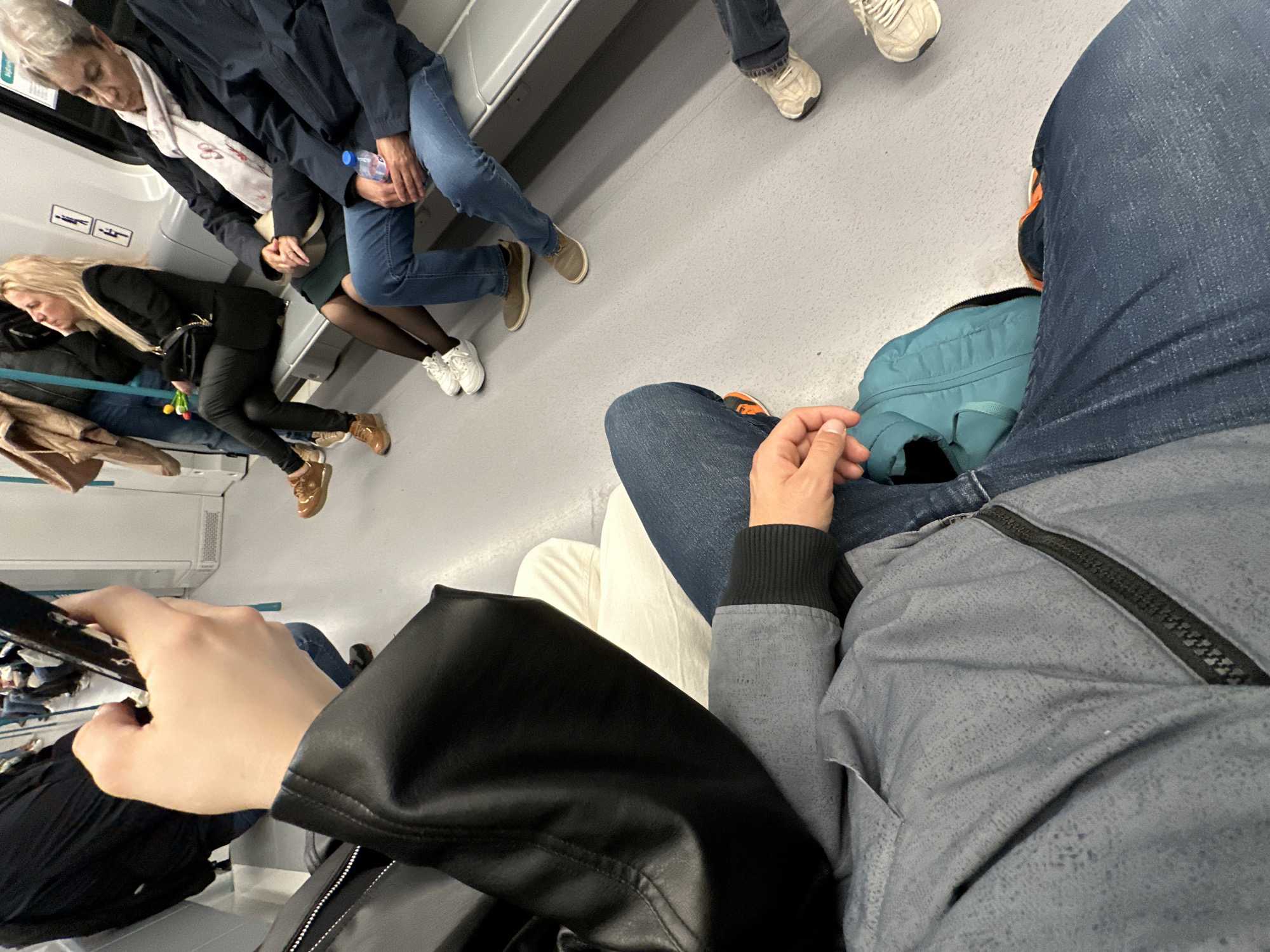 The image, taken from the subjective perspective of a passenger, depicts the interior of a public transportation vehicle, likely a subway train, in Sofia, Bulgaria. Several passengers are seated, with a visible group of four individuals. One individual is wearing a dark-colored coat over a white scarf, seated beside a person in blue jeans holding a water bottle. Across the aisle, another woman with blond hair is wearing a black jacket. Further down, there's a passenger wearing a light brown jacket. The floor is a light gray.
In the foreground, the photographer's hand is holding a phone, suggesting the image was captured with a mobile device. They are wearing a black leather jacket and a gray jacket with a zipper. A light blue bag is situated between their legs. The photo offers a candid snapshot of everyday urban life on the Sofia Metro.