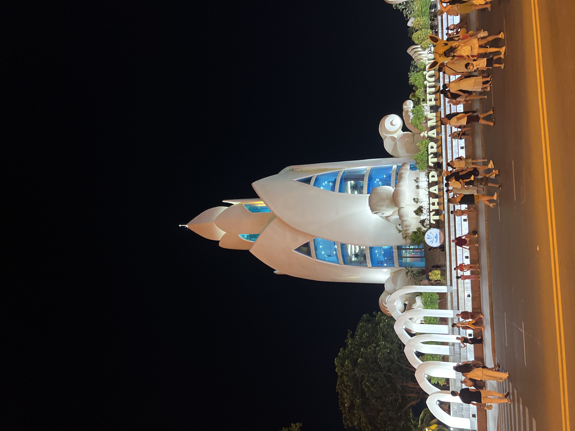 The image shows the Thap Tram Huong landmark in Nha Trang Ward, Vietnam, at night. The landmark is a tall, white, lotus-shaped building illuminated with blue lights. In front of the landmark, there is a large white archway structure. A group of people are walking across a street in front of the landmark, some are in pairs or small groups. The street is paved with asphalt and has yellow lane markings. There are trees and other vegetation visible to the left and behind the landmark. The sky is dark, indicating nighttime. The people are dressed in casual summer clothing. One person is wearing a Pikachu costume.