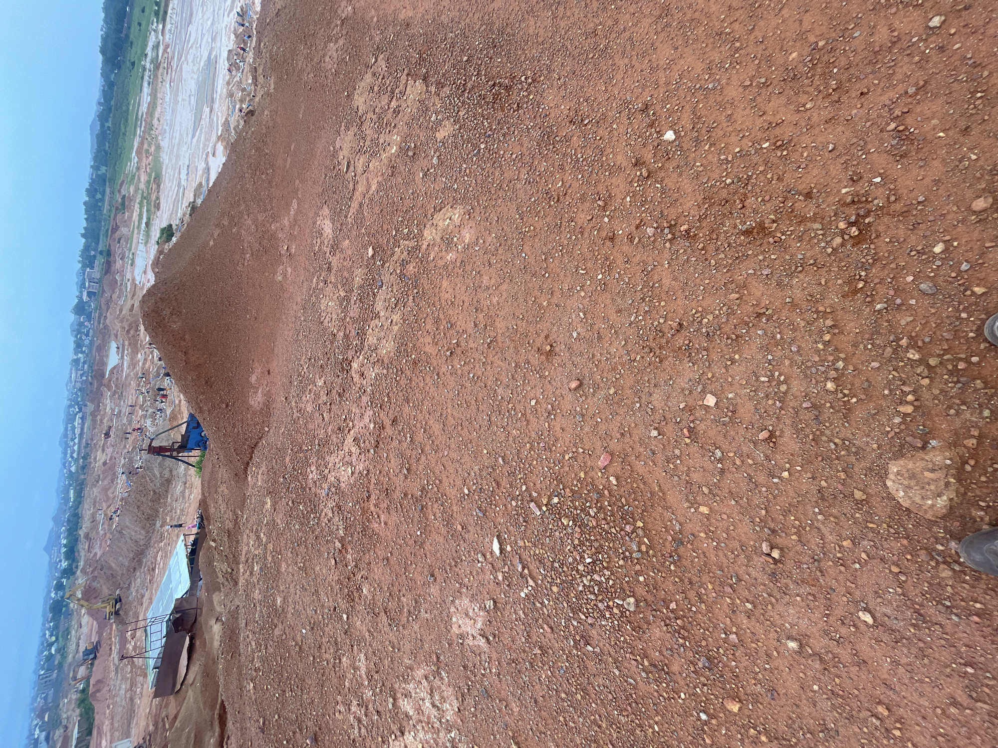 The image depicts an aerial view of a large-scale mining operation in Reifild-Ist, Nigeria. The terrain is predominantly reddish-brown soil with scattered rocks and gravel. In the foreground, the ground is uneven, with visible tire tracks suggesting the movement of heavy machinery.
Further back, the landscape reveals excavated areas, mounds of earth, and a body of water that appears to be a pit or pond. Several excavators are present, actively engaged in digging and moving soil. A temporary structure with a light-colored roof is also visible, possibly serving as a shelter or processing area.
Numerous individuals are scattered throughout the site, engaged in various tasks. Some appear to be working with the machinery, while others are seen in smaller groups, possibly manually sorting or collecting materials. The overall impression is one of an active industrial site in a somewhat arid or dusty environment. The sky is overcast, suggesting a diffused light condition.