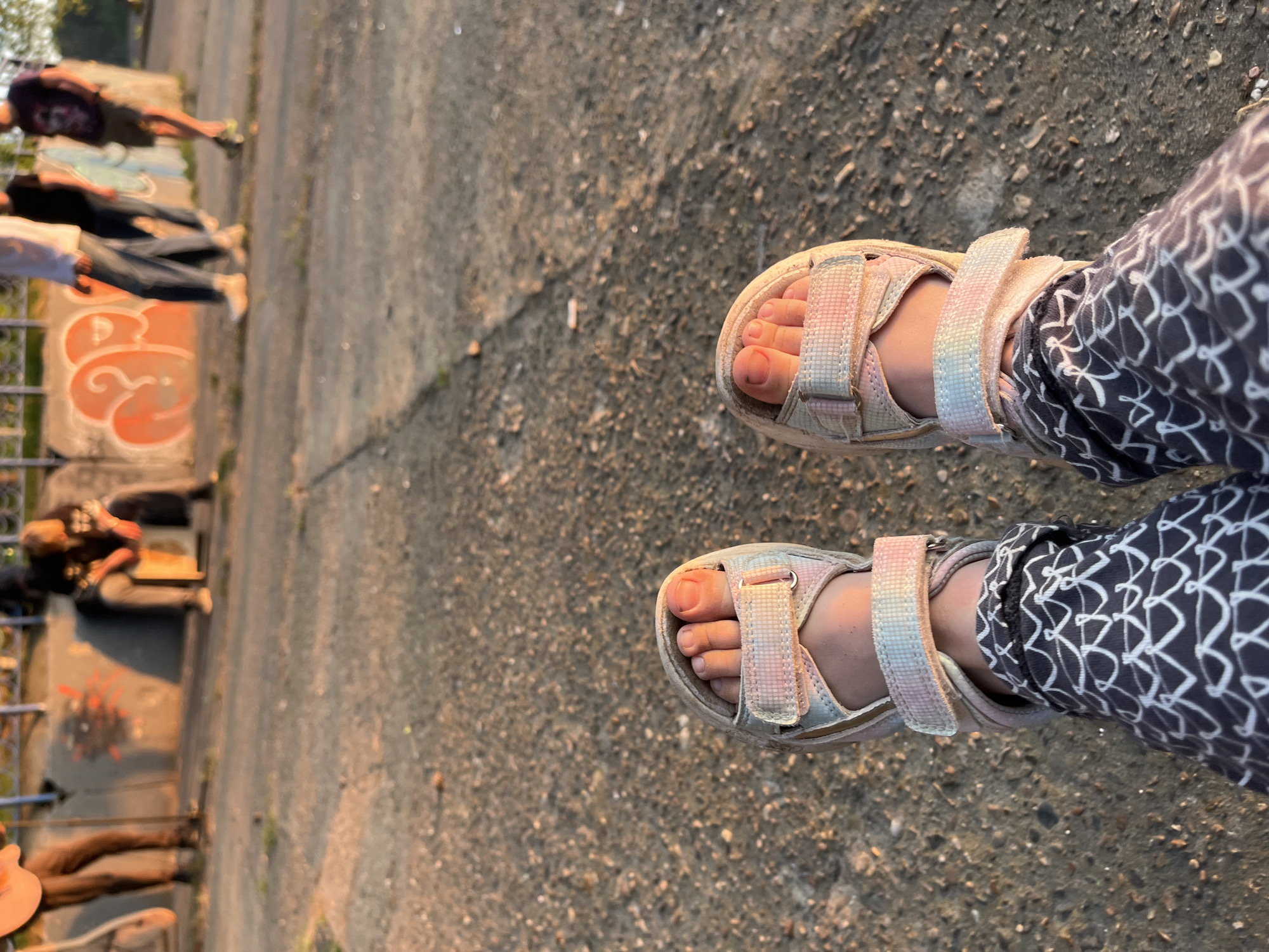 The image shows a pair of feet in sandals on a concrete surface. The person is wearing leggings with a white and dark gray pattern. In the background, there are several people sitting or lying down near a concrete structure with graffiti. The specific location in the city of Unknown, Unknown is not discernible from the image.