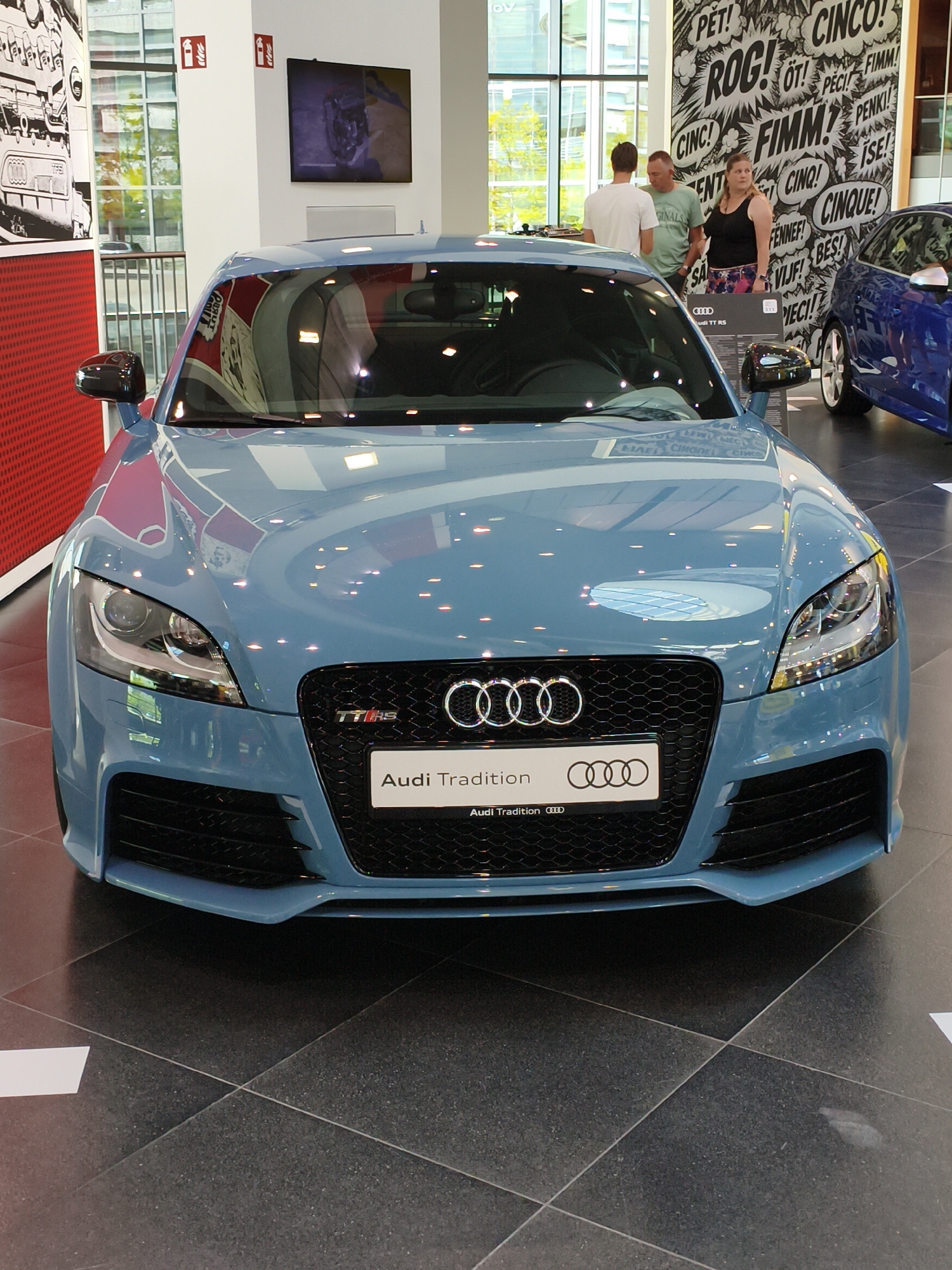 The image shows a light blue Audi TT RS parked indoors, likely in a showroom. The car is the central focus, with its front grille and headlights prominently displayed. The Audi logo and "Audi Tradition" are visible on a plate below the grille. In the background, there are several people, a blue Audi car, and a wall with a black and white graphic design featuring various words in speech bubbles. The floor is tiled with dark grey squares. The overall setting appears to be a modern exhibition space. The Audi Forum Ingolstadt is a likely location, given the prominent display of Audi vehicles and the surrounding context.