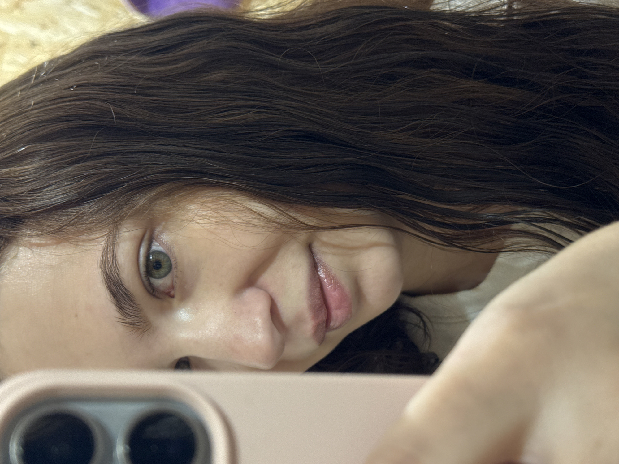 A close-up selfie of a person with dark brown, wet hair. The person has fair skin, green eyes, and a small smile. They are holding a pink phone in the foreground, partially obscuring their face. The background appears to be a wooden surface, possibly a ceiling or wall. There is nothing present to confirm the location as Nizhnyaya Gar', Russia.