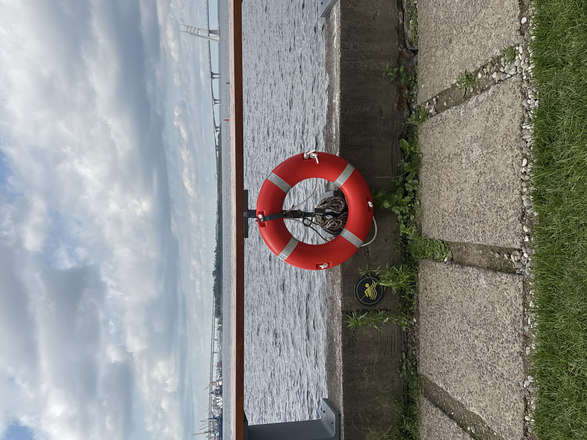 This is a photograph of a waterfront scene in Saint Petersburg, Russia, likely on the Neva River. In the foreground, a concrete embankment is visible, with patches of grass and paved stone pathways. A bright red life preserver with silver reflective stripes hangs from a metal fixture attached to a wooden railing that runs horizontally across the mid-ground of the image. The water of the river is choppy, reflecting the overcast, cloudy sky. In the background, a long, modern cable-stayed bridge stretches across the water. To the left of the bridge, a port with cranes and ships is visible. The overall atmosphere is somber and industrial.