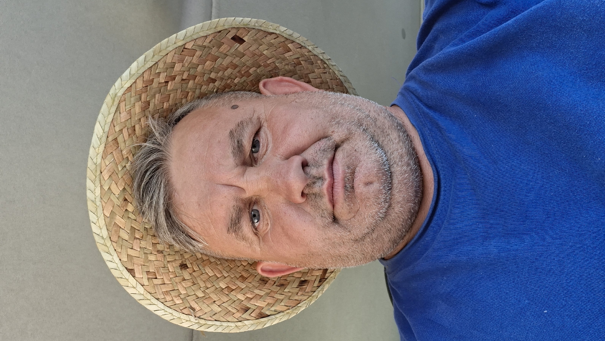 A low-angle, close-up, selfie-style photograph shows a middle-aged man wearing a woven straw hat and a blue t-shirt. The man has fair skin, graying hair, and a stubbly beard. His expression is neutral, with his eyes looking directly at the camera. He has a mole on his right cheek. The background is a plain, light-colored surface, possibly a ceiling or wall, which could be found in a residential or commercial interior in Hîncești, Moldova. The lighting appears to be natural daylight coming from above.