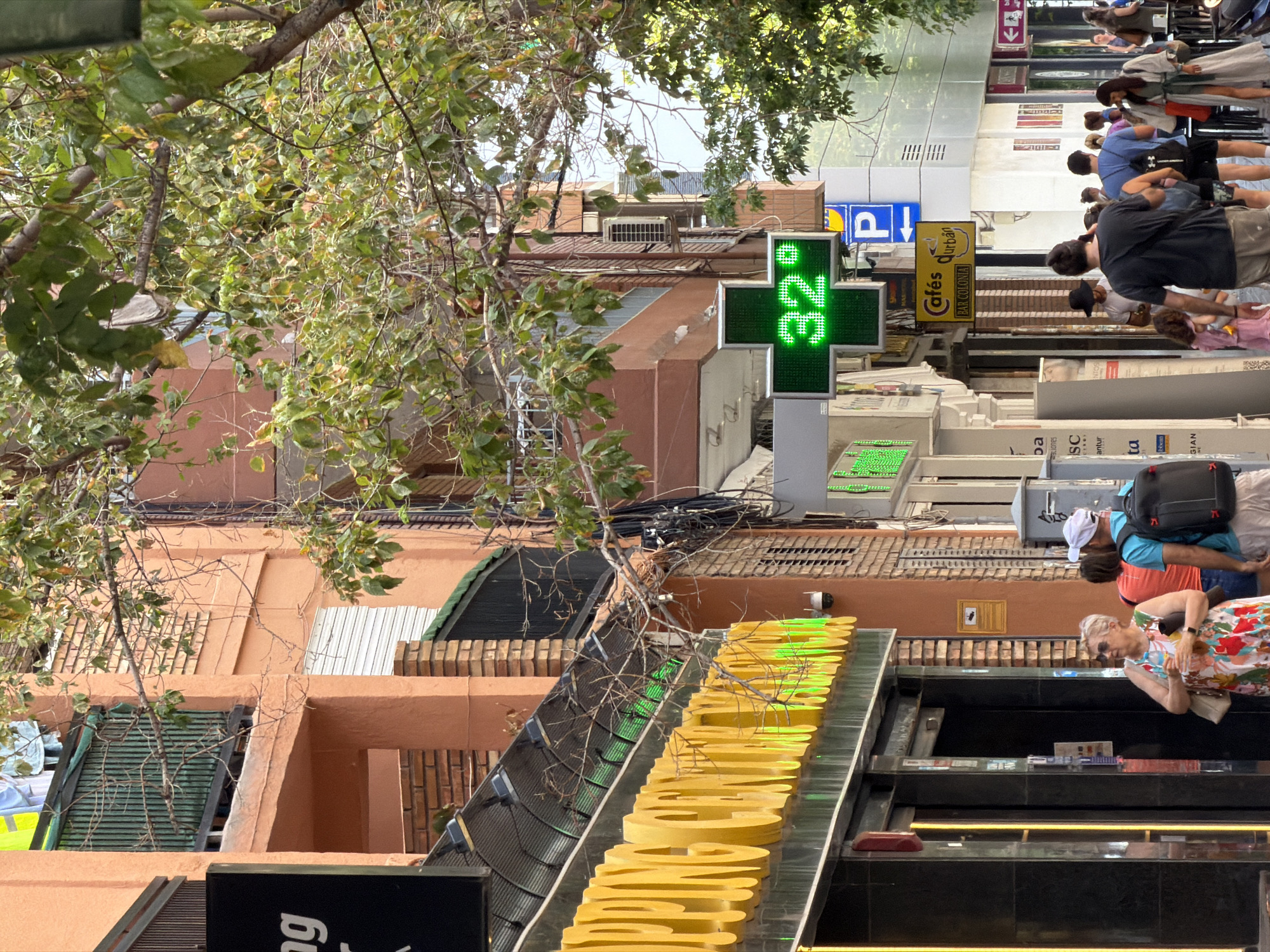This media file depicts a street scene with several people walking and standing. A pharmacy sign with a green cross and a digital display showing "32°" is prominently visible. There are also signs for "Cafés durban" and "BAR COLONIA". The architecture suggests an urban environment, possibly in a warm climate given the foliage and the high temperature displayed. The lighting suggests it is daytime. The people are casually dressed, and their expressions are neutral, suggesting a normal day. Some are carrying bags, indicating they might be shopping or commuting. The overall impression is a bustling street with commercial activity.