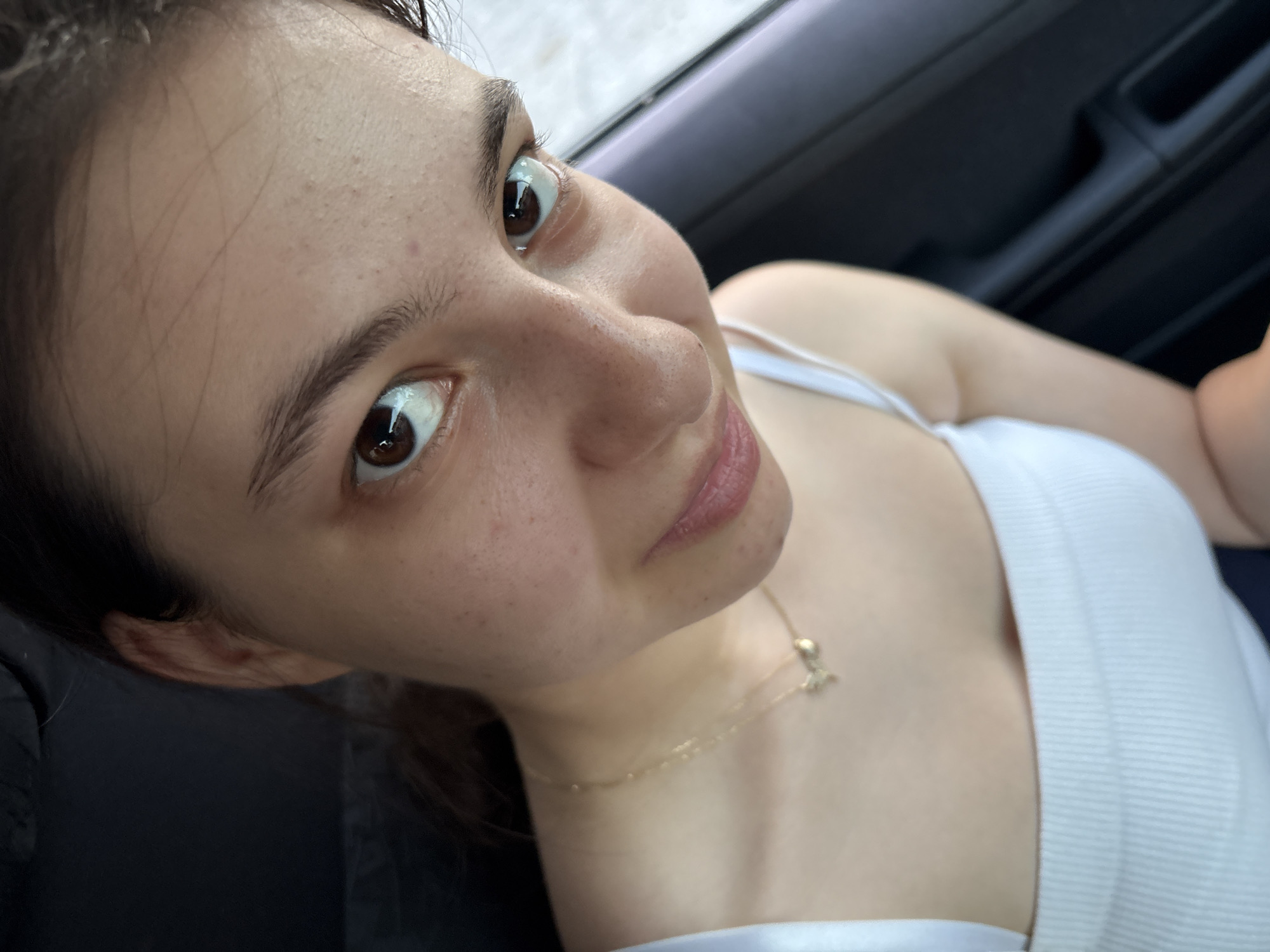 A close-up, high-angle shot features a young woman with brown eyes and dark hair, looking directly at the camera. She is wearing a white ribbed tank top and a delicate gold necklace with a small bow pendant. The background is dark, suggesting an indoor setting or a car interior, with the right side of the frame showing a dark surface with vertical lines, possibly a car door panel. The lighting is natural and highlights the woman's facial features, including light freckles on her cheeks and nose. There are no overt indications of the location being in Tervel, Bulgaria, or any specific urban environment.
