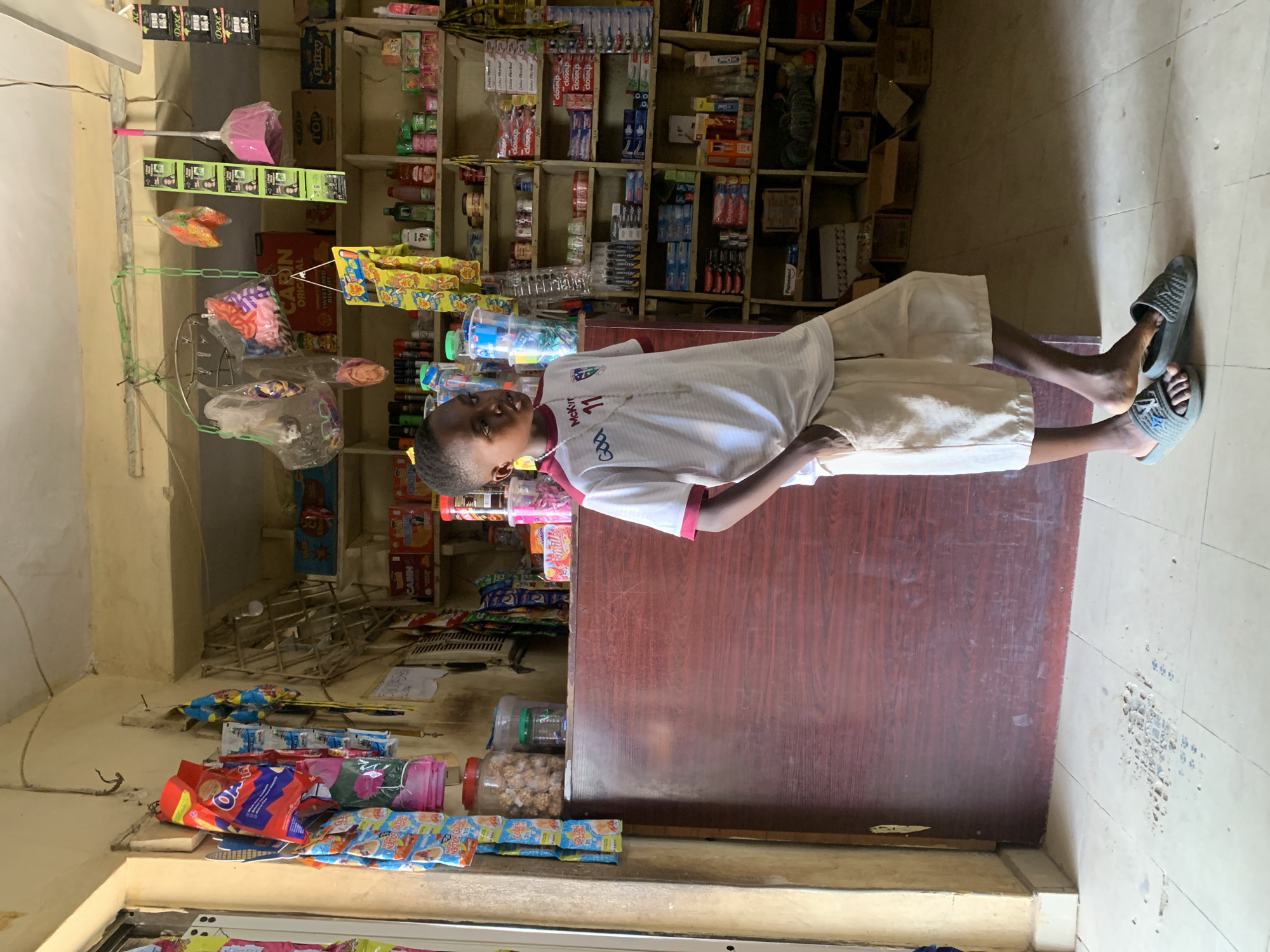 A full-shot photograph captures a young boy standing in what appears to be a small, local convenience store or kiosk in Adankolo, Nigeria. The boy, of African descent, stands in the foreground, facing slightly towards the viewer with a neutral expression. He is wearing a white short-sleeved polo shirt with maroon trim on the collar and sleeves, featuring a logo that includes "McKvr" and "God 11". He has light-colored shorts and grey slip-on sandals on his feet.
The store's interior is functional and well-stocked. Behind the boy, a dark wooden counter runs horizontally, separating him from the densely packed shelves lining the back wall. These shelves are made of light-colored wood and hold a wide variety of consumer goods. Visible items include numerous packaged snacks, bottled drinks, and personal care products such as toothbrushes (brands like "closeup," "Colgate," "Oral B"), toothpaste, and hair care items ("Dexe"). Some items, like small toys or candies, are hung in plastic bags from a makeshift display structure suspended from the ceiling. A pink broom is also hanging overhead. On the floor in the foreground, near the bottom left, are more packaged snacks.
The lighting suggests it is daytime, with light entering the room from an unseen source. The overall impression is that of a bustling, essential local business.
Visible text and brands include: "CABIN ORIGINAL," "TOP," "Spicy," "McKvr," "God 11," "Dosep," "closeup," "Oral B," "Colgate," and "Dexe."